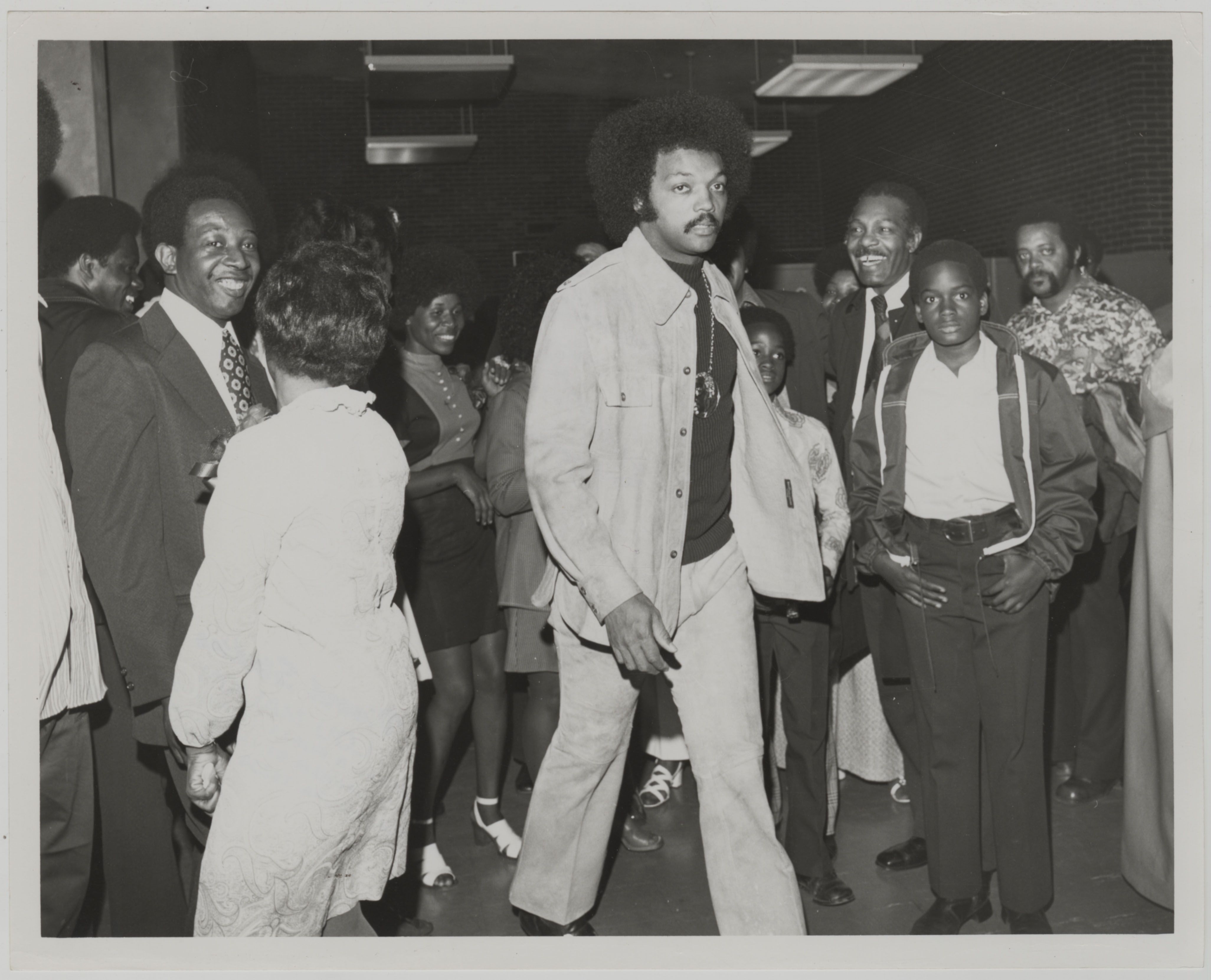 Black-and-white photo of a group of African American people indoors, dressed in 1970s style, engaging socially; man in center wears light jacket and pants with large afro.