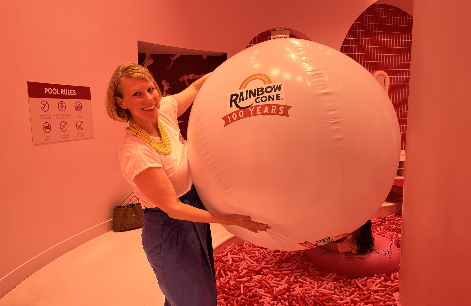 Smiling woman in a white top and yellow necklace hugs a giant inflatable pink ball reading "Rainbow Cone 100 Years" in a room with a "Pool Rules" sign and a floor covered in red candy pieces.