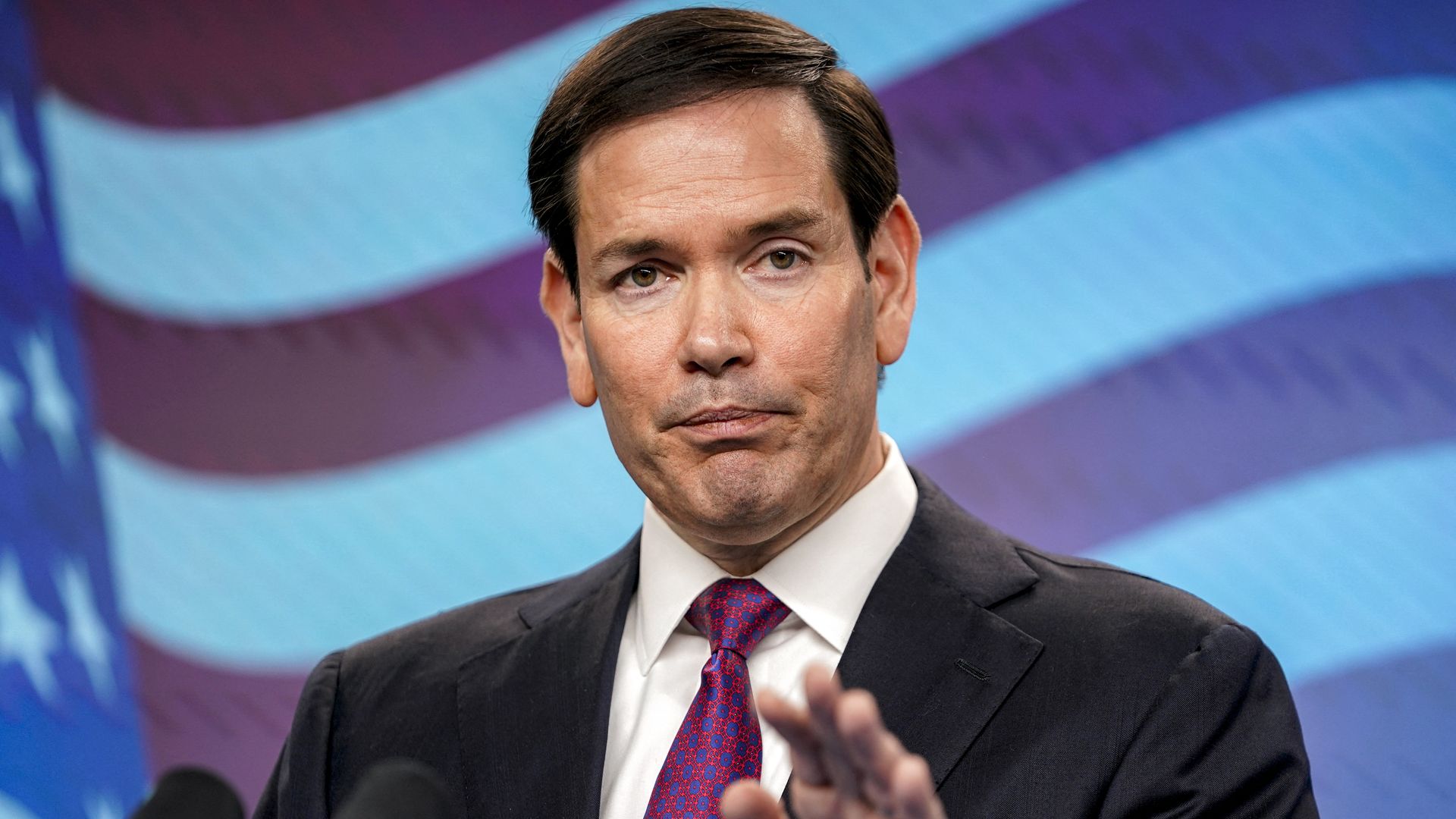 A black-haired Secretary of State Marco Rubio, wearing a black jacket, white shirt and pink tie with blue dots, purses his lips and gestures during a press conference in front of a TV screen displaying the US flag.