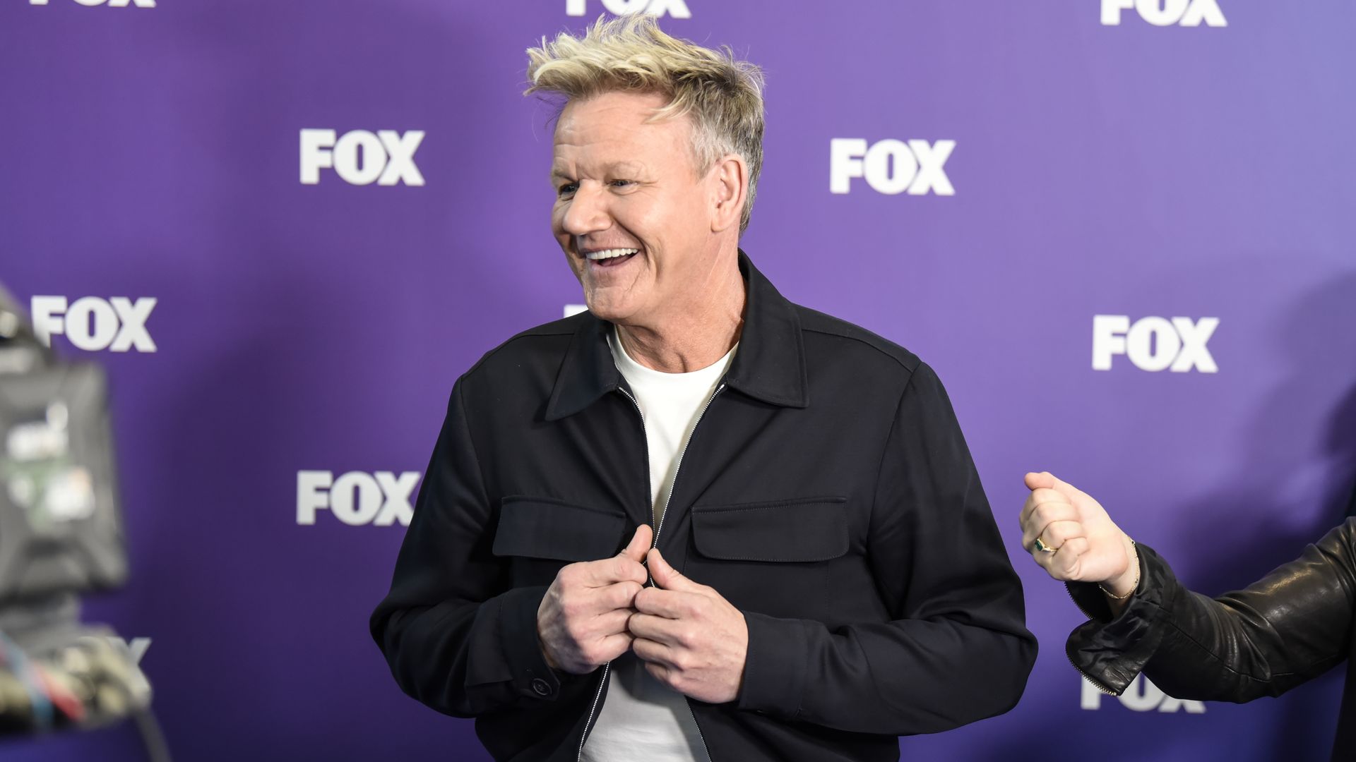 Gordon Ramsay stands on a red carpet and smiles as he holds the edges of his blazer jacket.