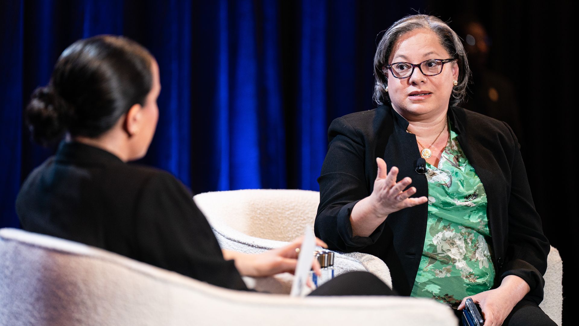 Two women sit in white armchairs on a stage with a blue curtain. The woman on the right wears a black blazer over a green floral blouse, glasses, and a microphone, gesturing as she speaks.