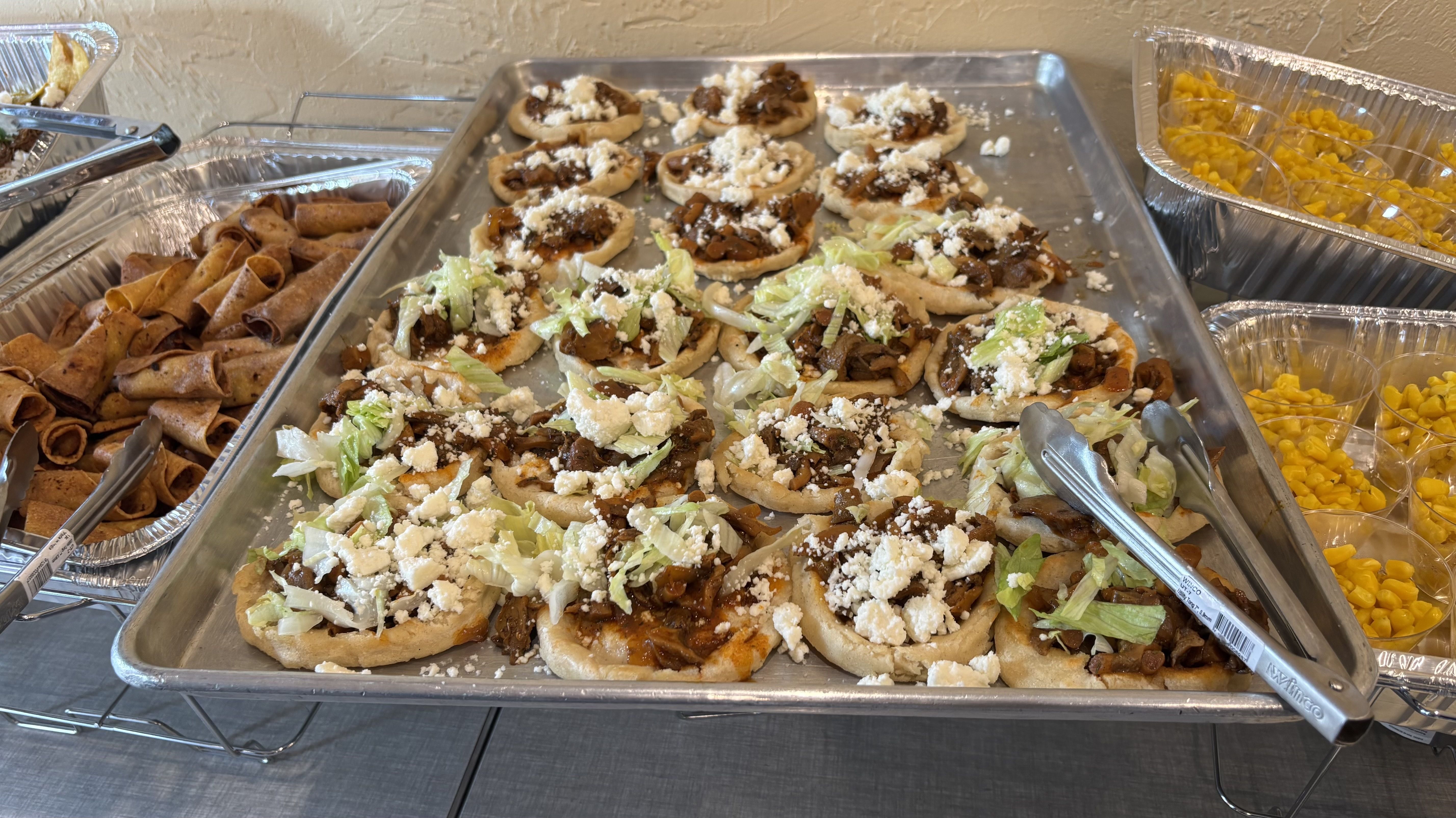 Buffet tray with bite-sized flatbreads topped with shredded lettuce, crumbled cheese, and sautéed mushrooms; nearby are rolled tortillas and yellow diced fruit in aluminum trays.