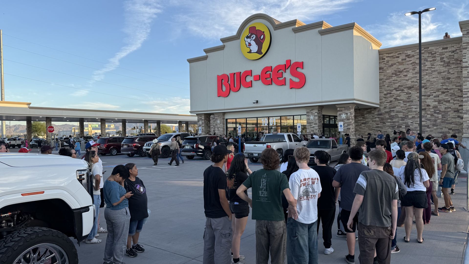 Wide exterior view of a Buc-ee's with a yellow circular logo with a buck-toothed beaver wearing a red cap, and large red letters; a large crowd gathers outside the entrance as cars fill the parking lot on a sunny day.