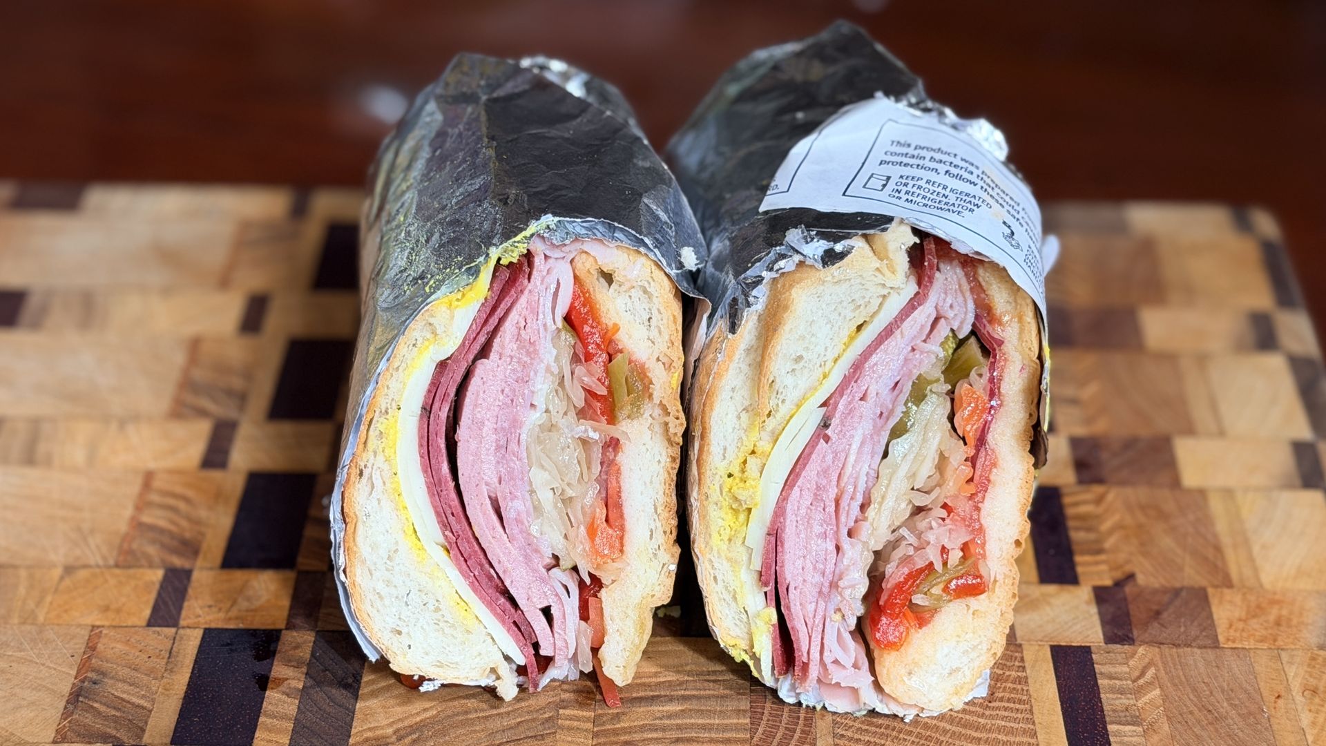 Two halves of a large deli wrap, showing layers of ham, cheese, peppers, onions, and bread, wrapped in foil with a label, on a wooden cutting board.
