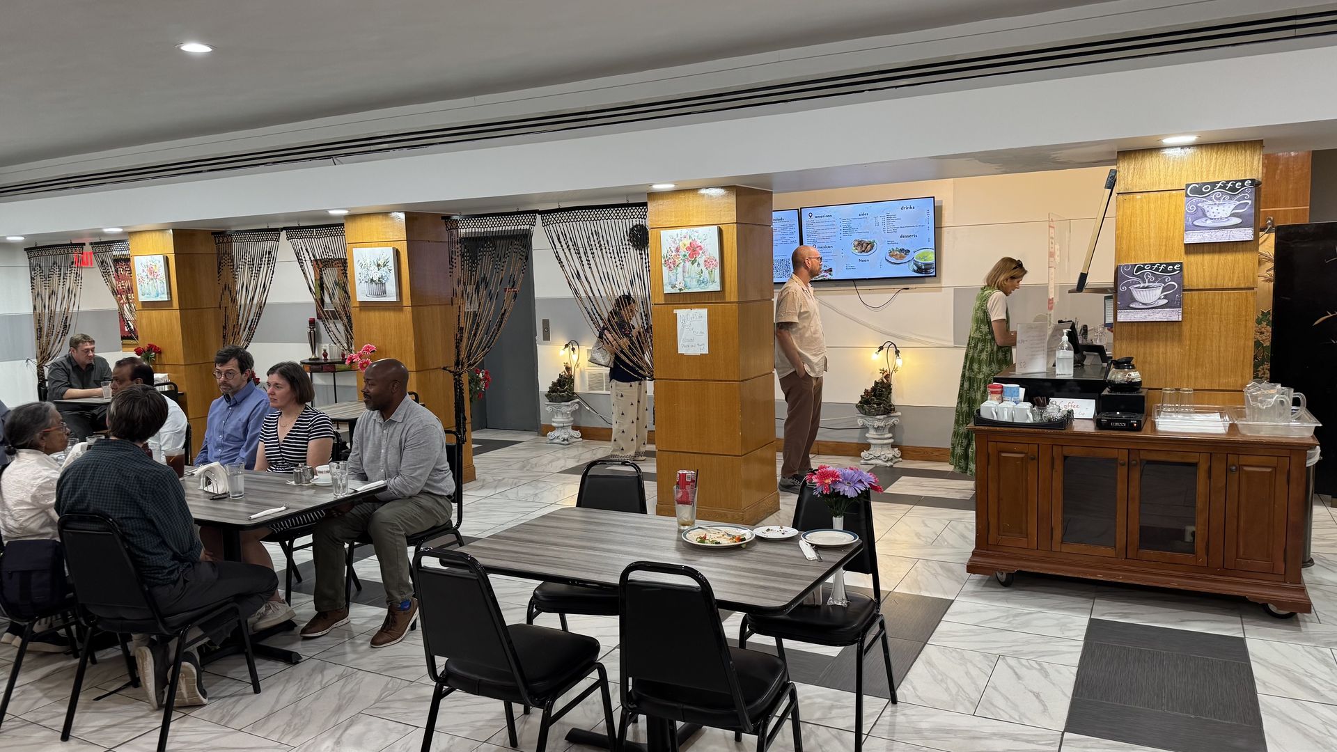 Busy dining area in a bright hall with several tables of patrons. A man in a light shirt stands by a digital menu screen; a coffee counter with chalkboard signs sits to the right, framed by wooden columns.