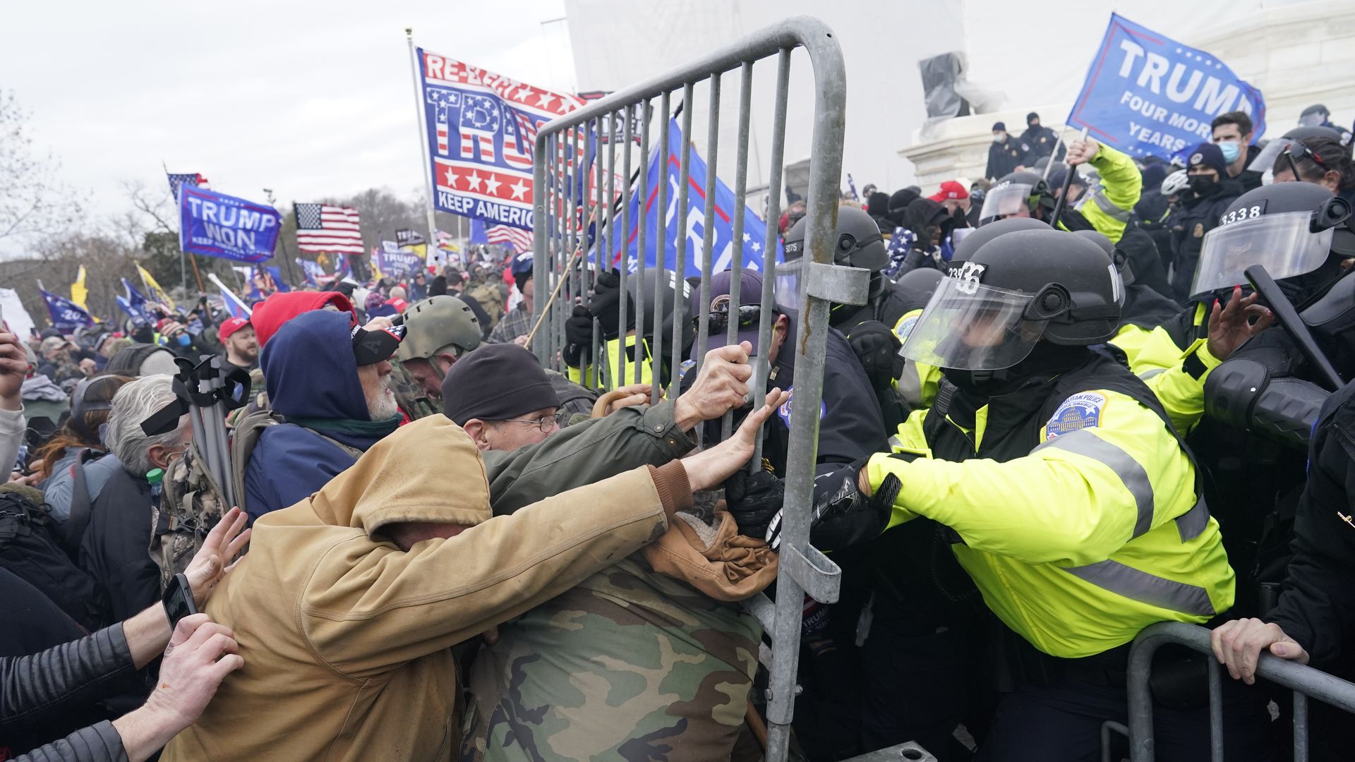 A yellow-vested police officer pushes against a mob of people with Trump flags in the background