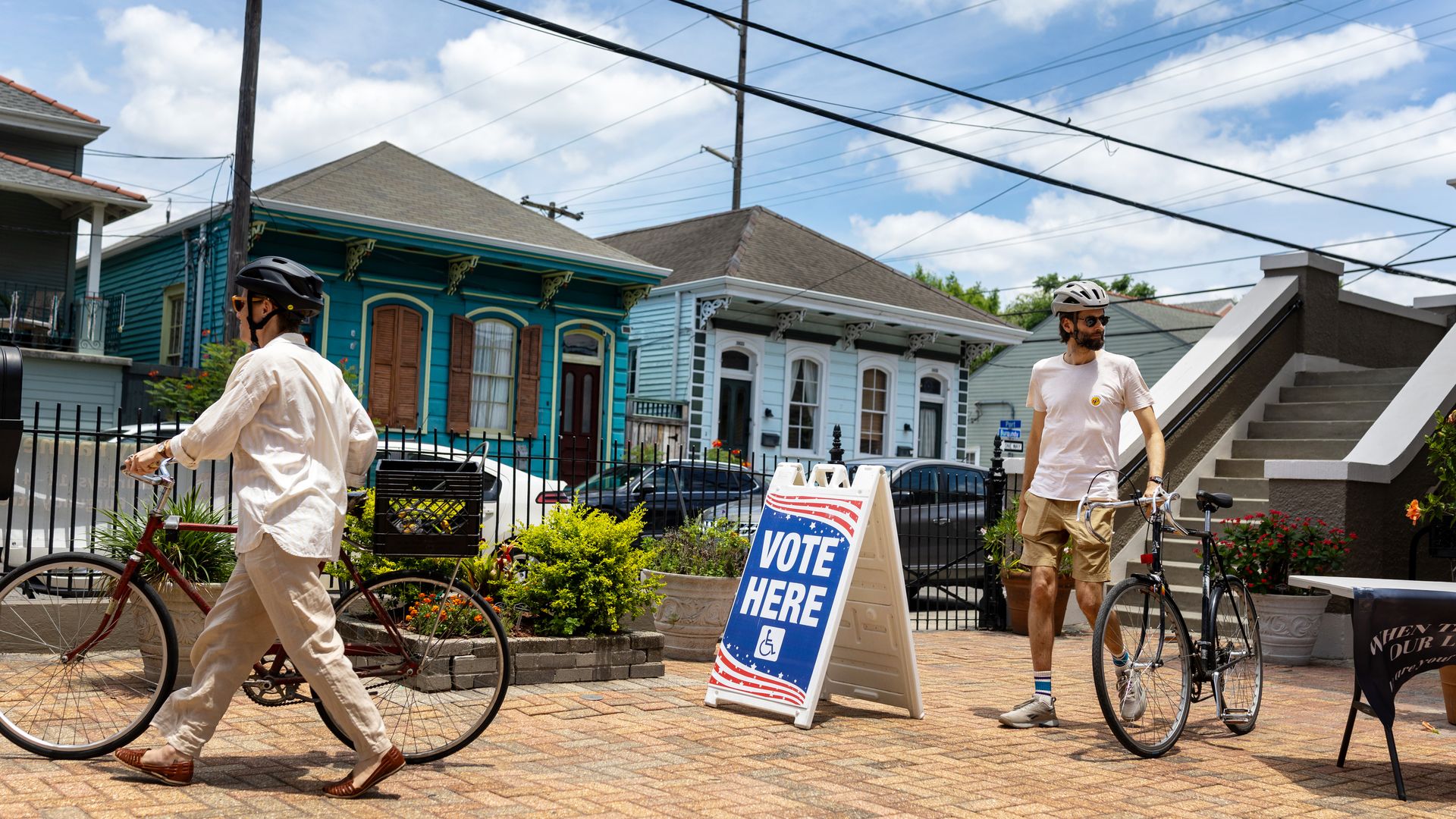 Two people wear bike helmets as they walk their bikes past a large sandwich board-style sign that says "vote here." Behind them are New Orleans shotgun homes.