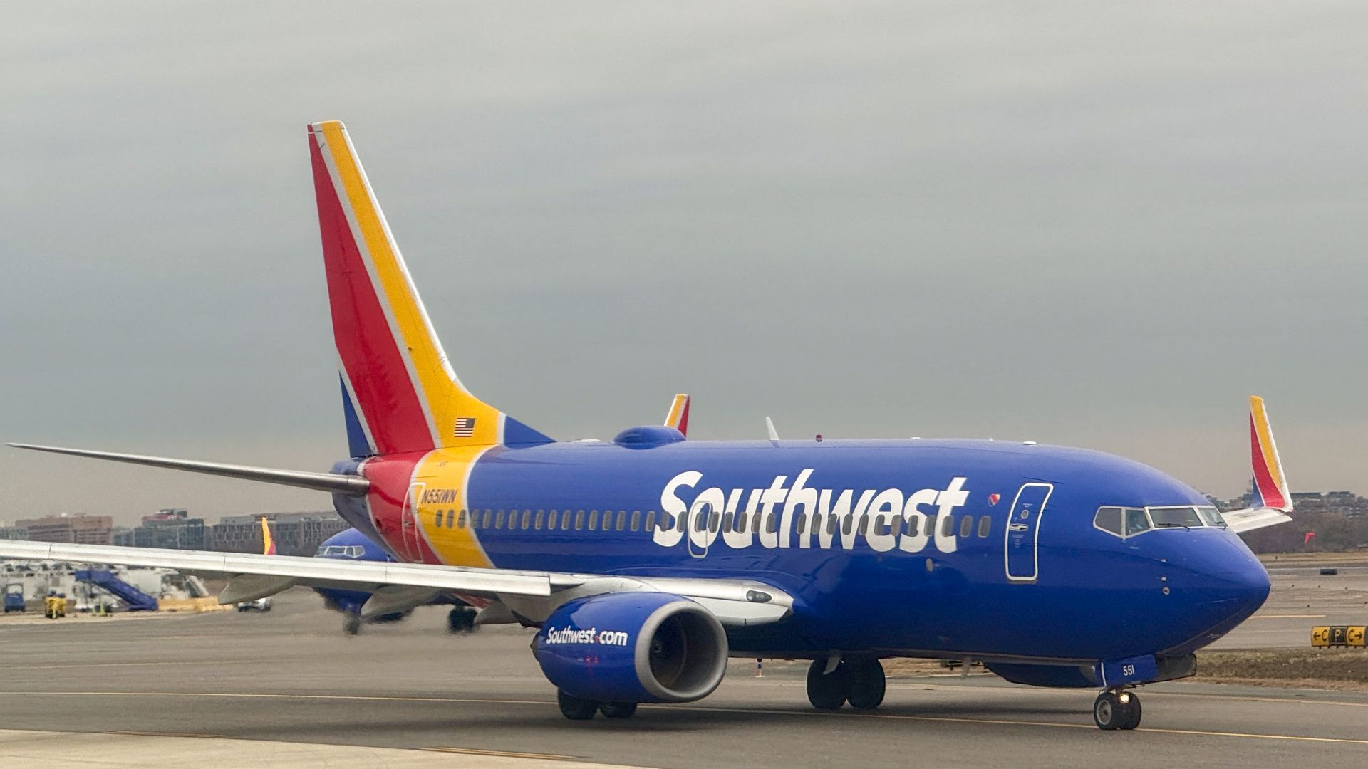 Blue Southwest Airlines airplane with yellow and red tail taxiing on a gray runway under an overcast sky at an airport.