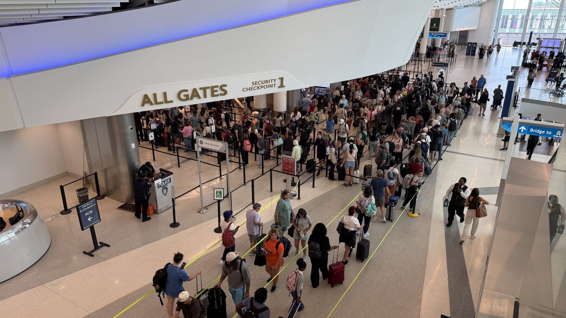 Busy airport terminal with a long security checkpoint queue beneath a white wall that reads "ALL GATES" and "Security Checkpoint 1". Passengers with luggage wait behind stanchions near glass walls.