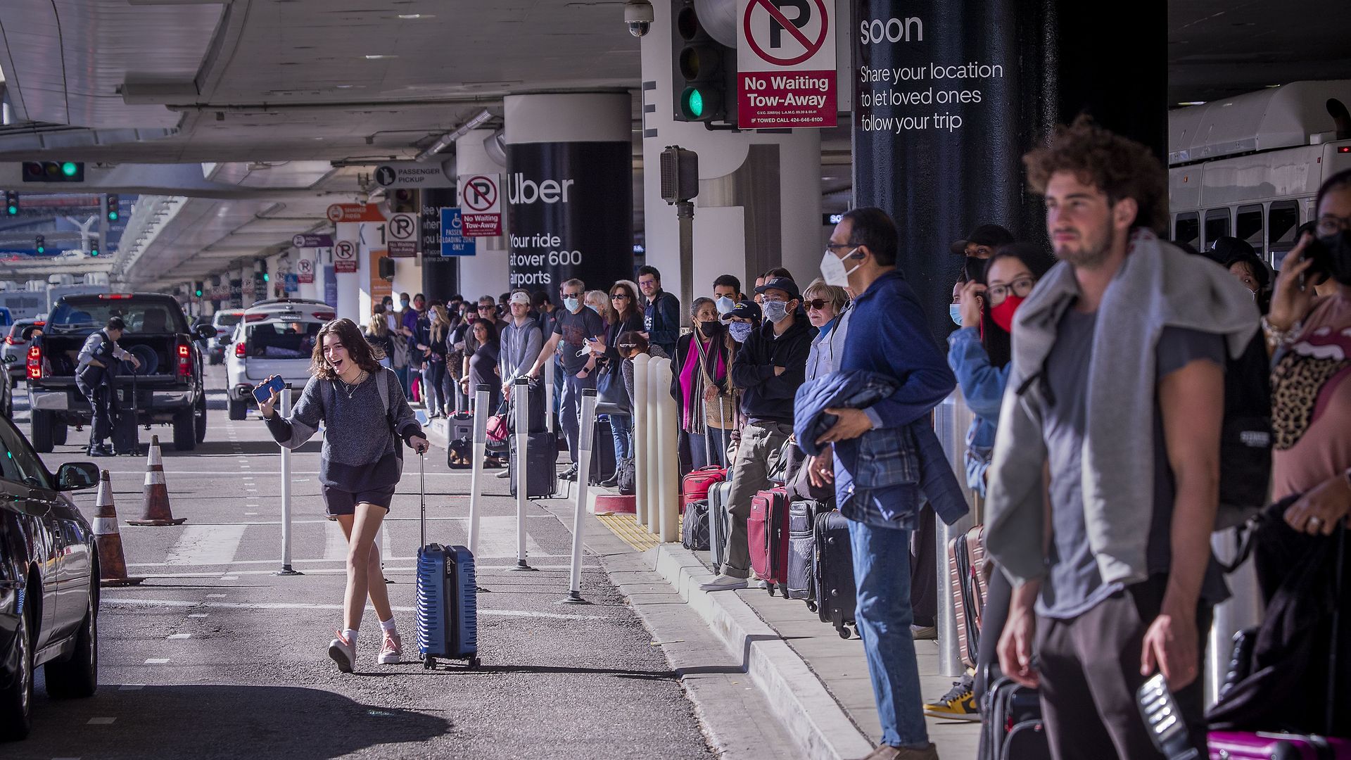 a crowd of people stand on a curb to the left, in the middle of a frame, a woman waves to a driver as she walks towards a vehicle. 