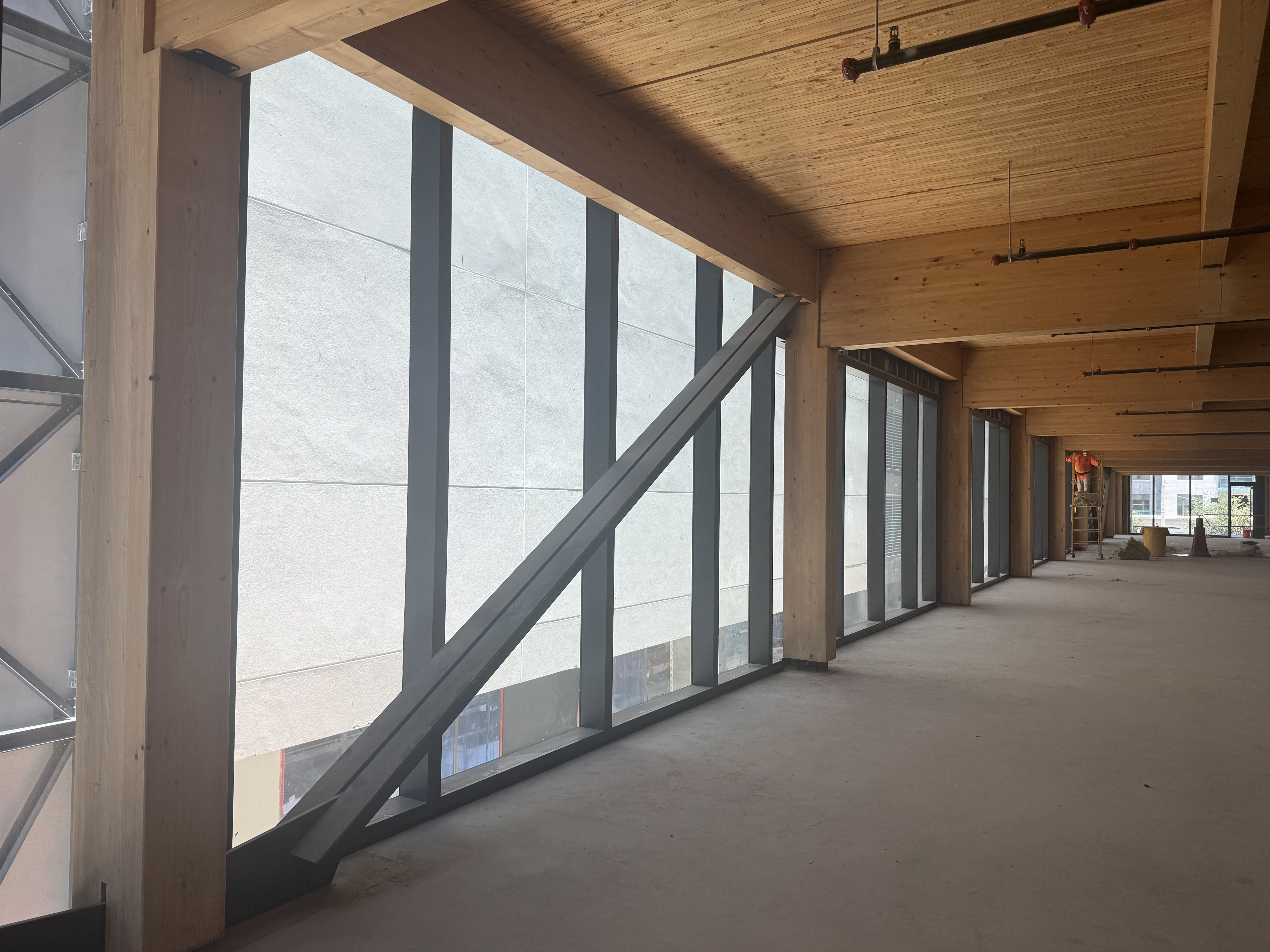 Indoor construction site with tall wooden beams, a wood-paneled ceiling, and large left-side windows. Exposed pipes overhead and a distant worker near orange cones.