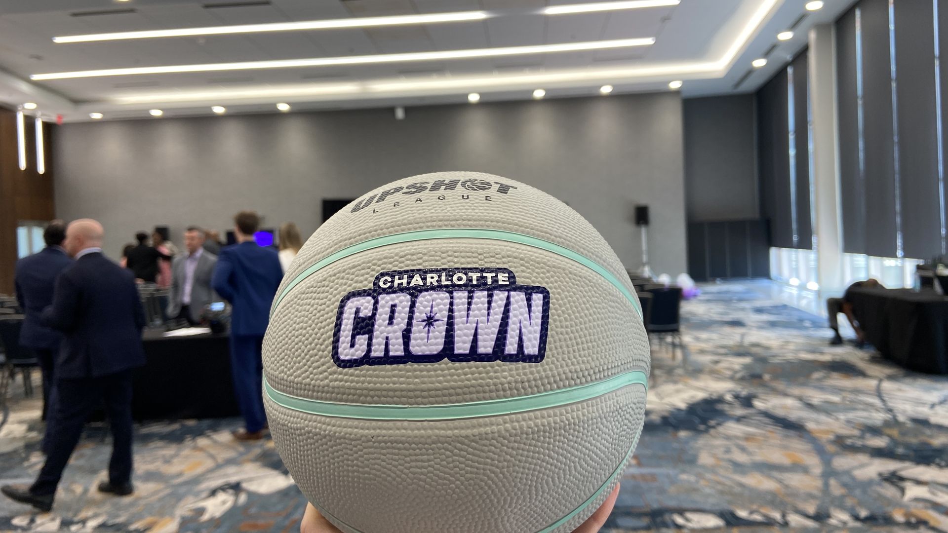 Close-up of a white UpShot League basketball with light blue stripes and "CHARLOTTE CROWN" logo held in a conference room with people in business attire blurred in the background.