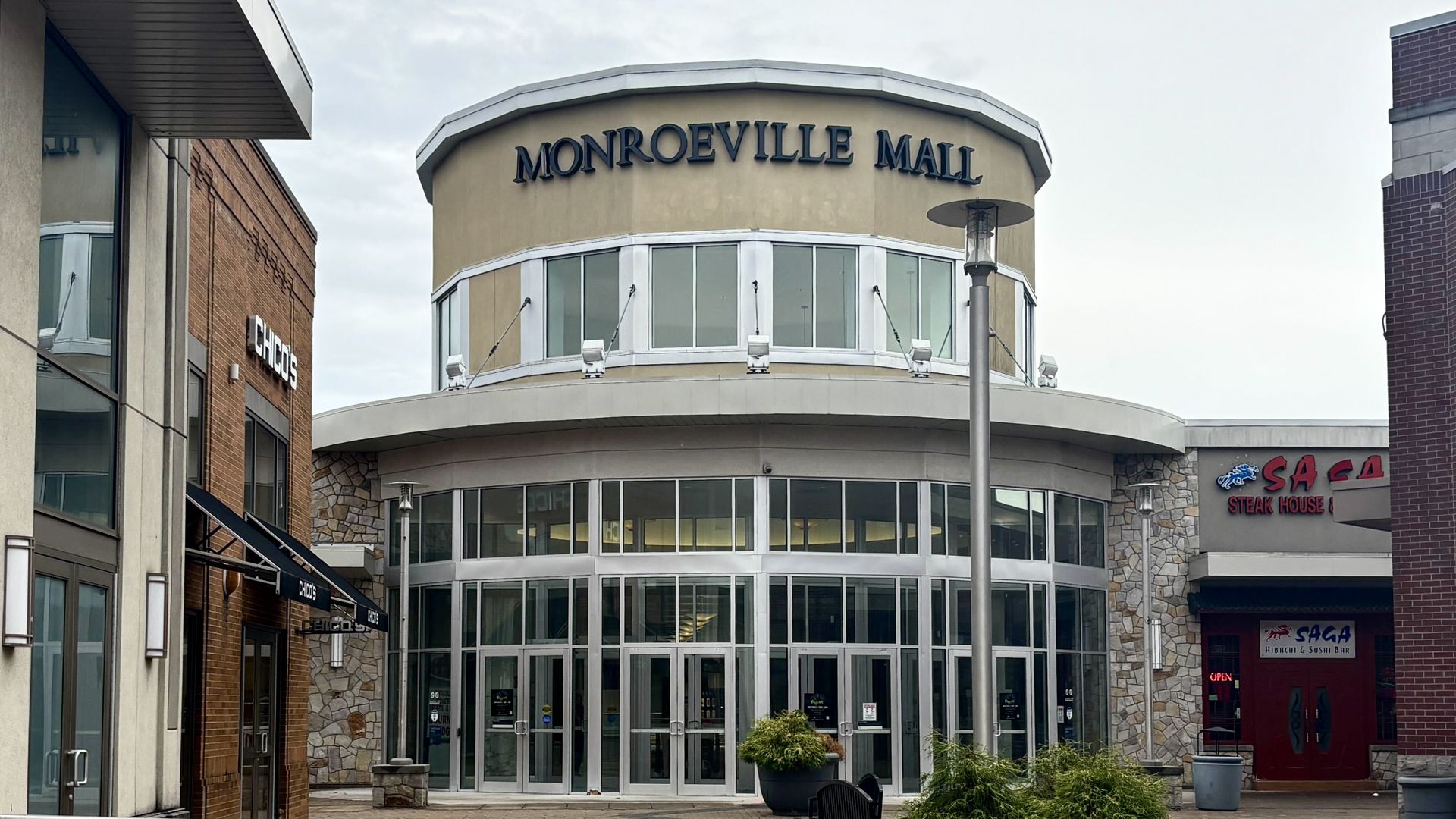 Entrance of Monroeville Mall with large glass doors and beige upper section. On left is Chico's store with brick exterior; on right is Saga Steak House and Sushi Bar with red door.