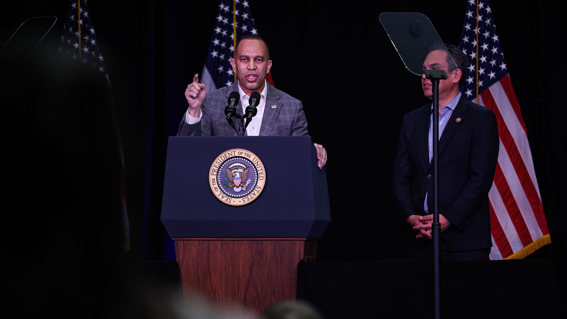 House Minority Leader Hakeem Jeffries, wearing a dark gray plaid suit and white shirt, speaks at a podium with the seal of the president in front of an American flag.