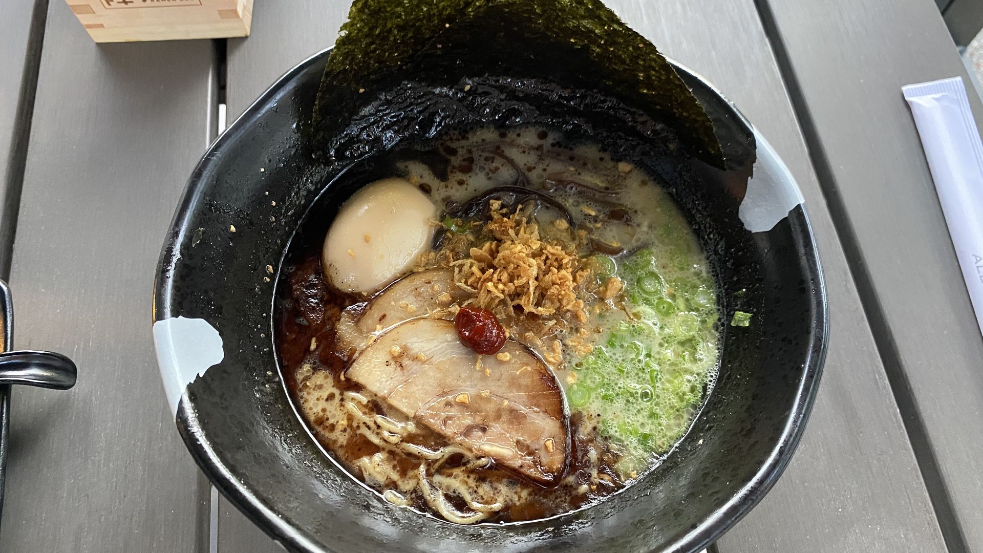 A black bowl filled with dark broth, noodles, pork slices, green onions and a hard boiled egg. 