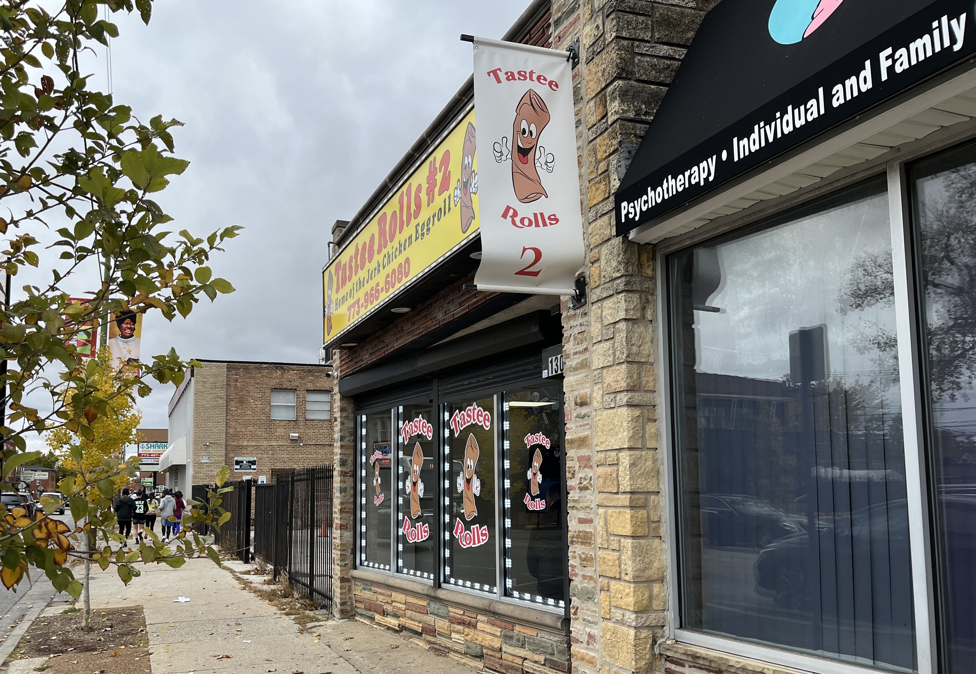 Street view of a storefront with a yellow sign and a banner advertising "Tastee Rolls 2," featuring a cartoon chicken eggroll giving thumbs up, next to a psychotherapy center.