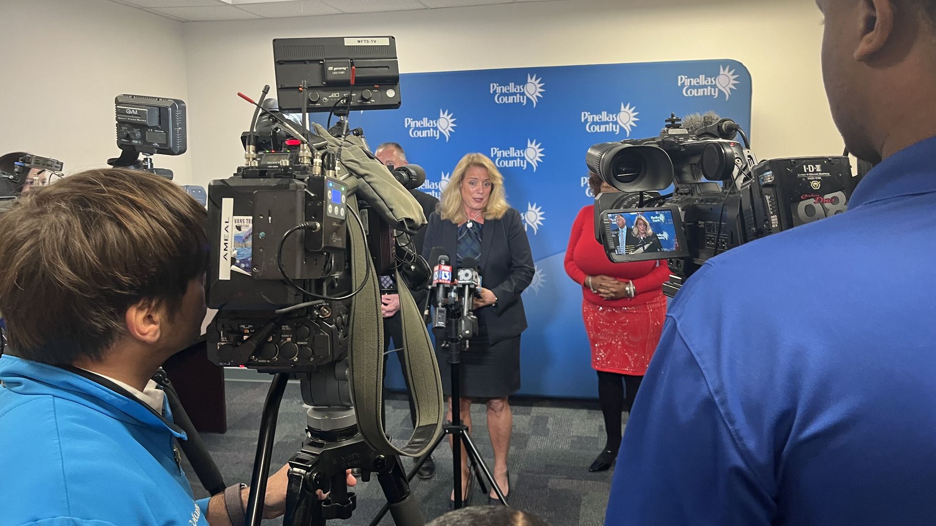 A woman in a black suit addresses several TV cameras while standing in front of a blue wall that says "Pinellas County." 