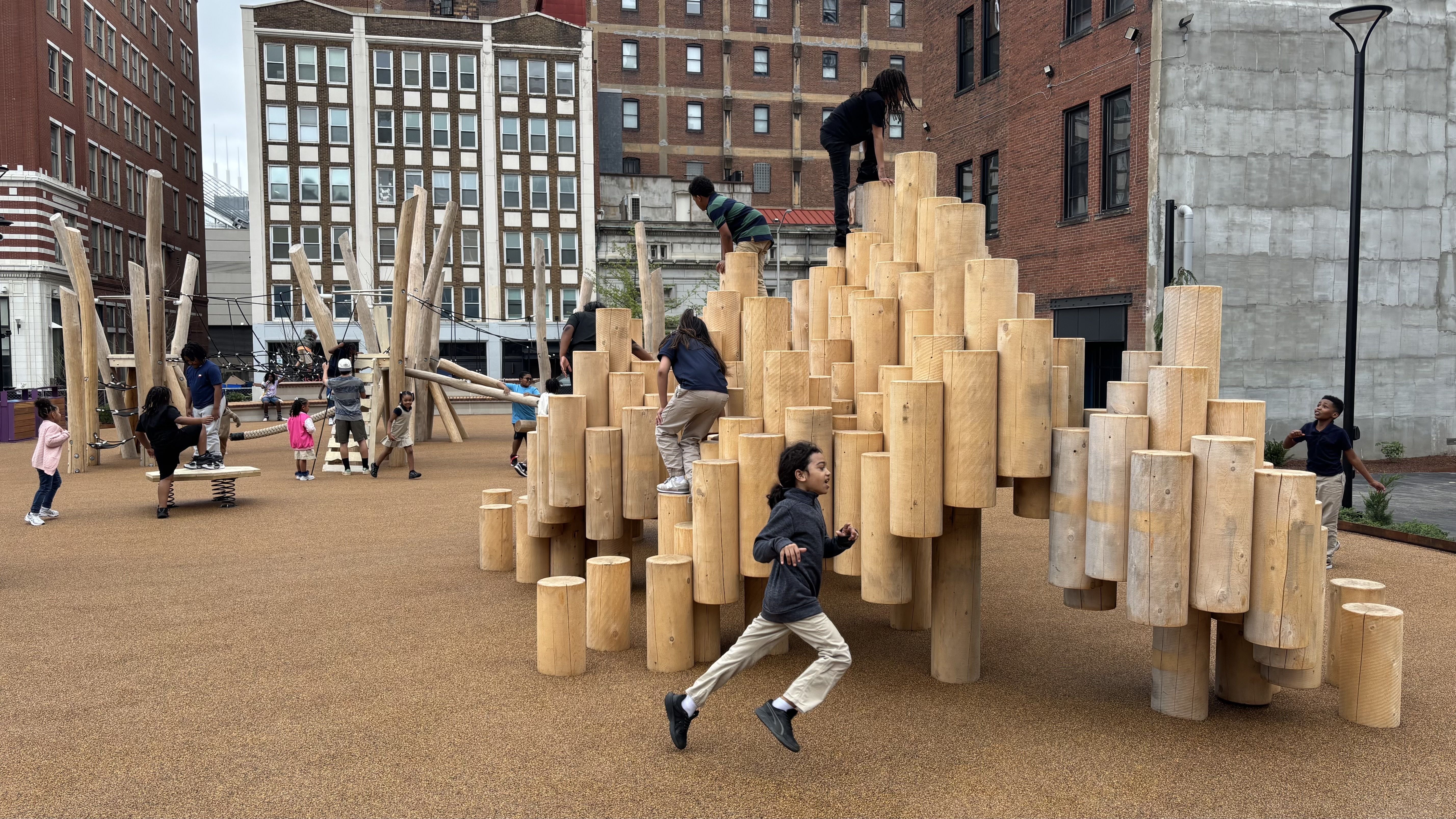 Urban playground featuring a mound of tan, stacked wooden logs for climbing; children climb and run on the logs as brick buildings rise in the background on brown rubberized ground.