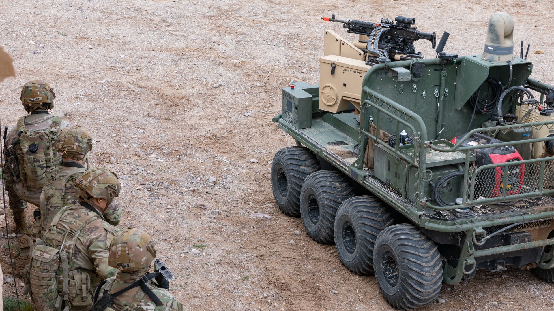 U.S. Army soldiers stack against a wall on the left while a military transport vehicle moves up on the right side. They are standing in dirt and sand.