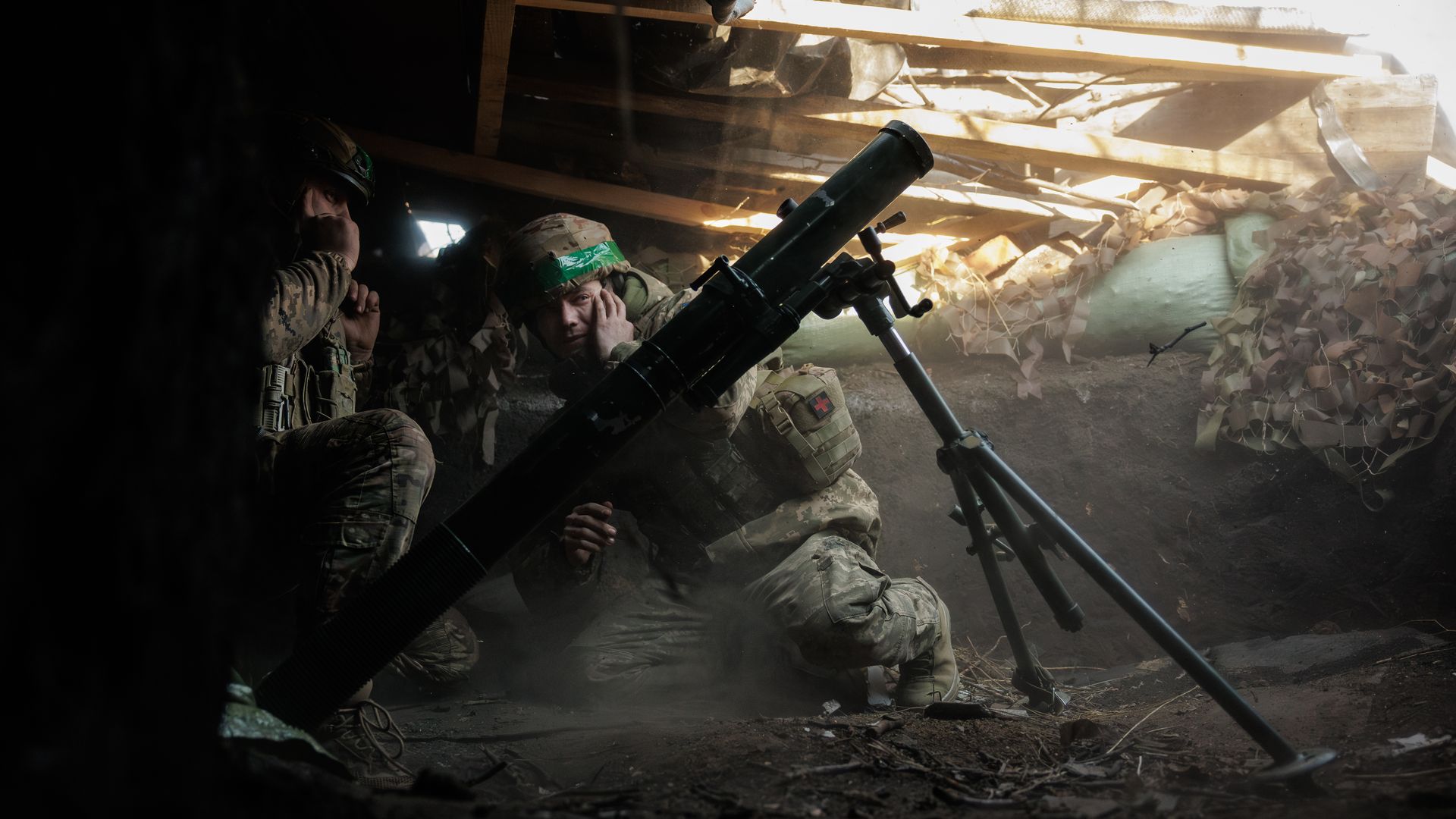 Two soldiers in camouflage uniforms and helmets crouch in a dark, dusty bunker with a large mortar weapon, shielding their ears, sunlight filtering through wooden slats above.