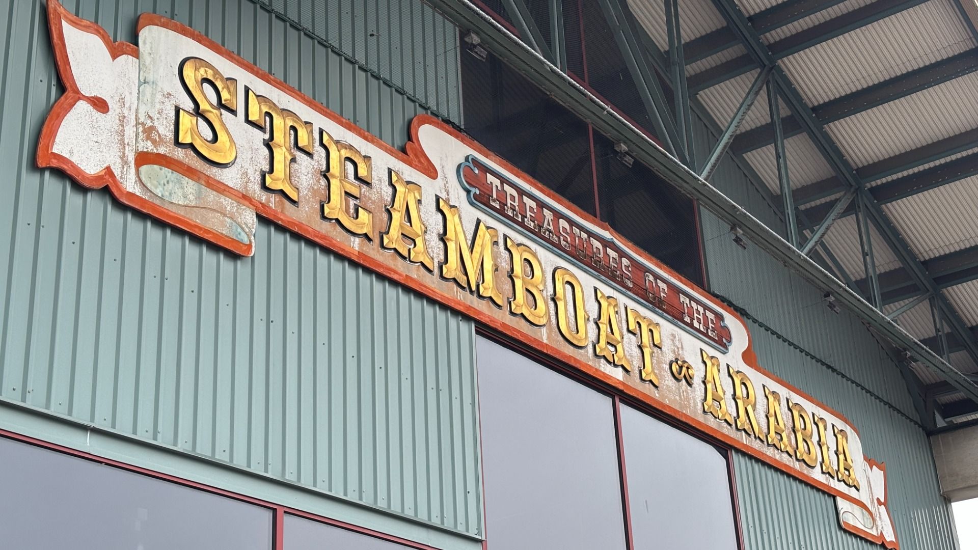 Weathered vintage sign reading "STEAMBOAT" in large gold letters on a green corrugated metal wall, with red trim; smaller "TREASURES OF THE" text above and steel roof trusses overhead.