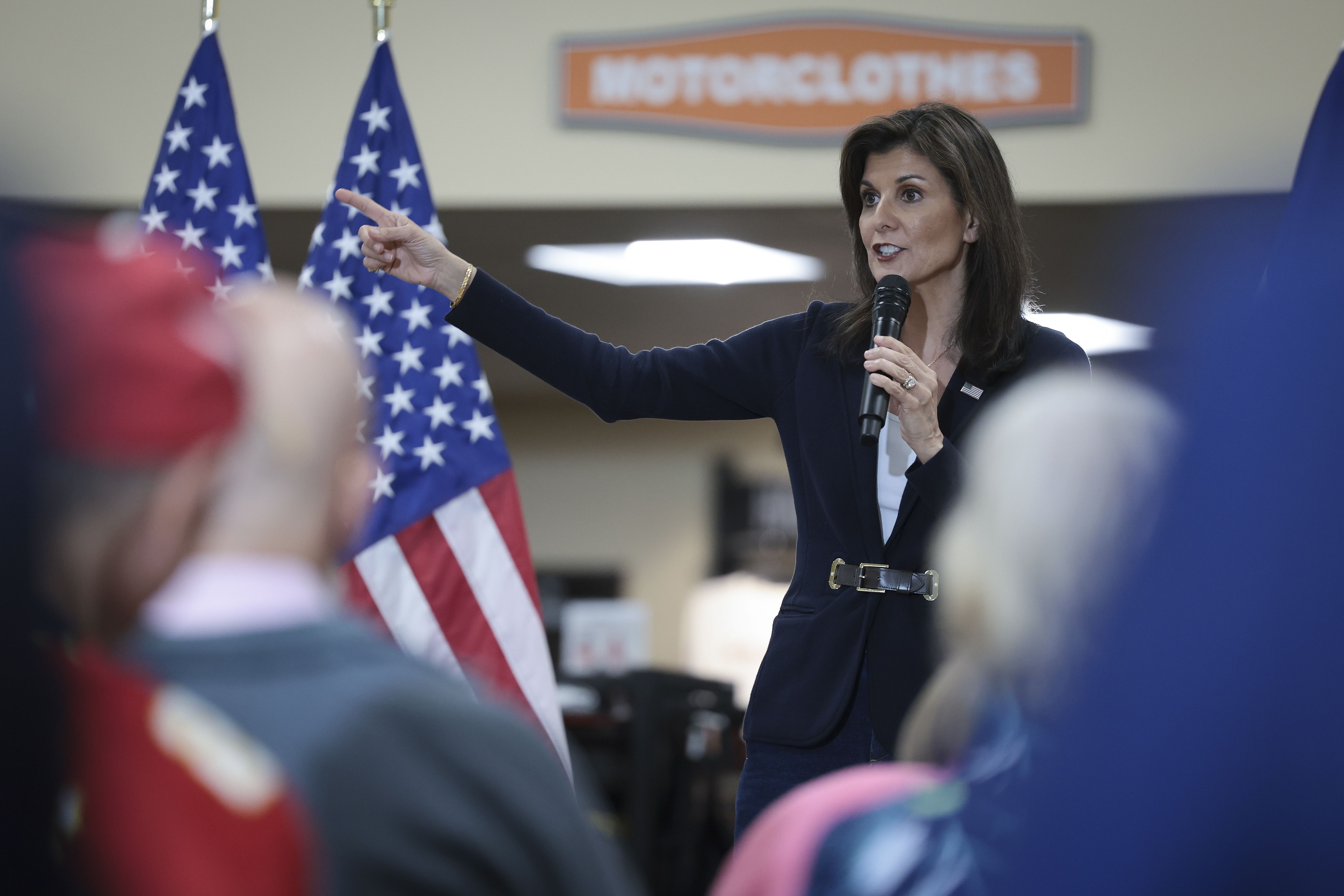 Nikki Haley in a black jacket and belt speaking into a microphone with flags to her side