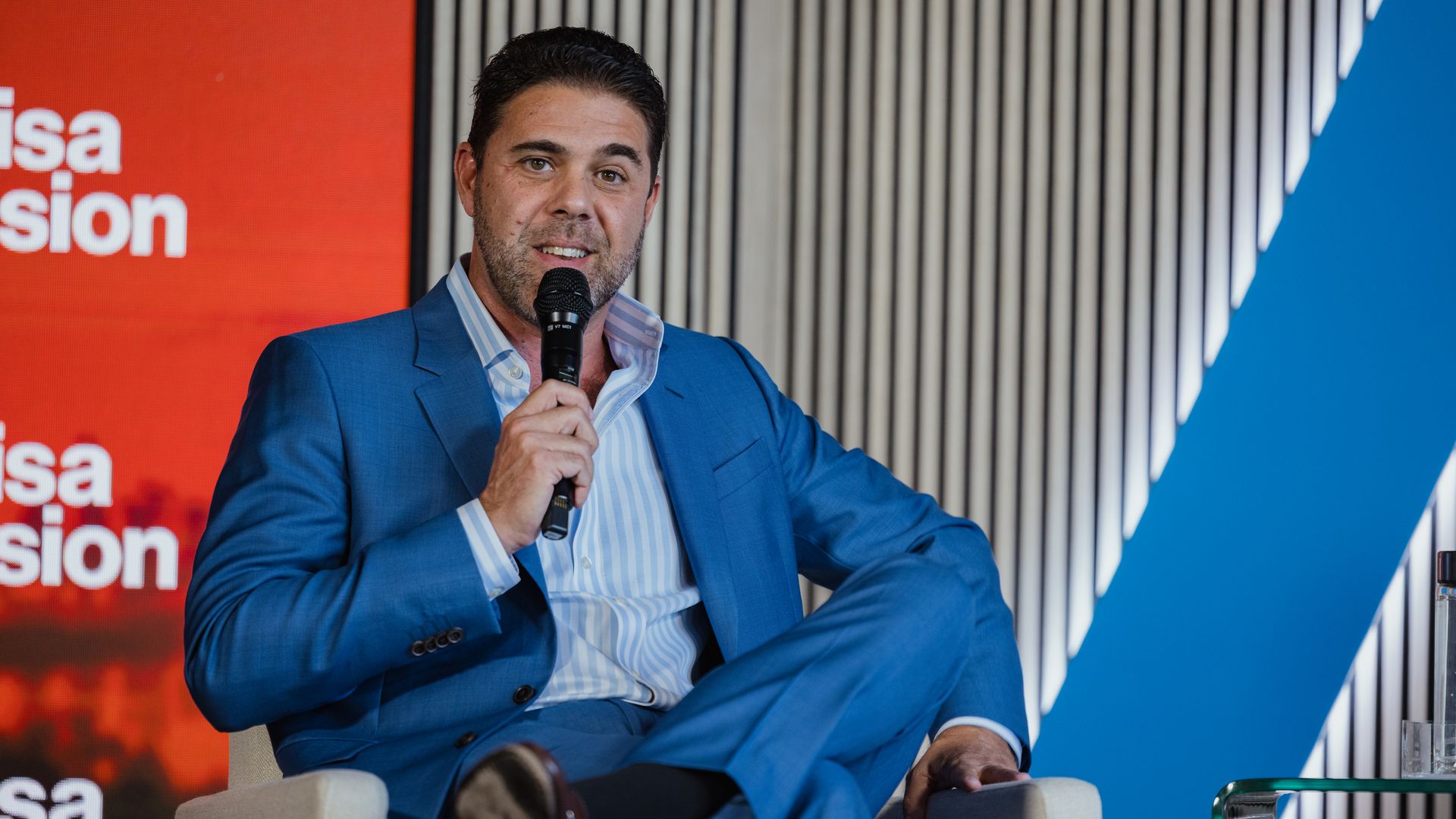 A man in a blue suit sits on a beige chair, holding a microphone and speaking. Behind him is a red panel with white text and a striped wall.