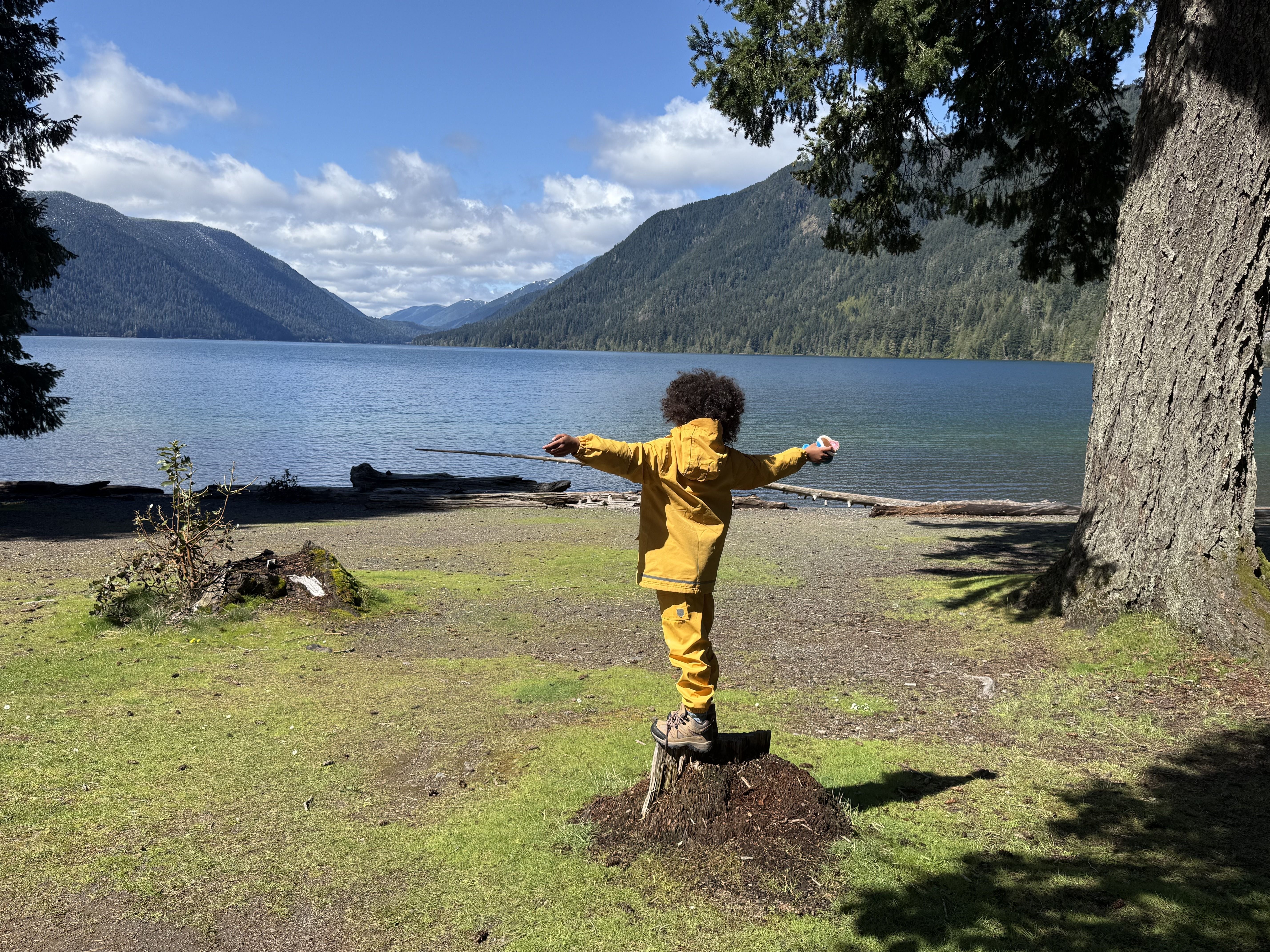 A little boy in a yellow rainsuit stands on a log with his arms spread in front of a blue lake.