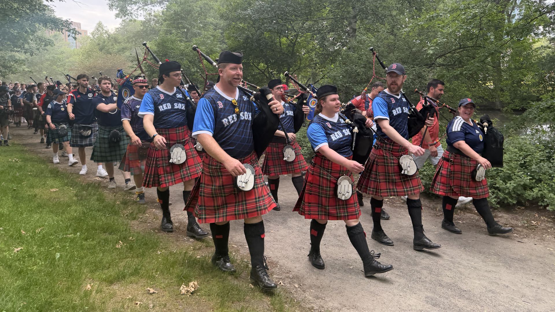 Parade of bagpipers in red plaid kilts and blue jerseys marching along a dirt path in a leafy park, carrying bagpipes and smiling as spectators line the route.