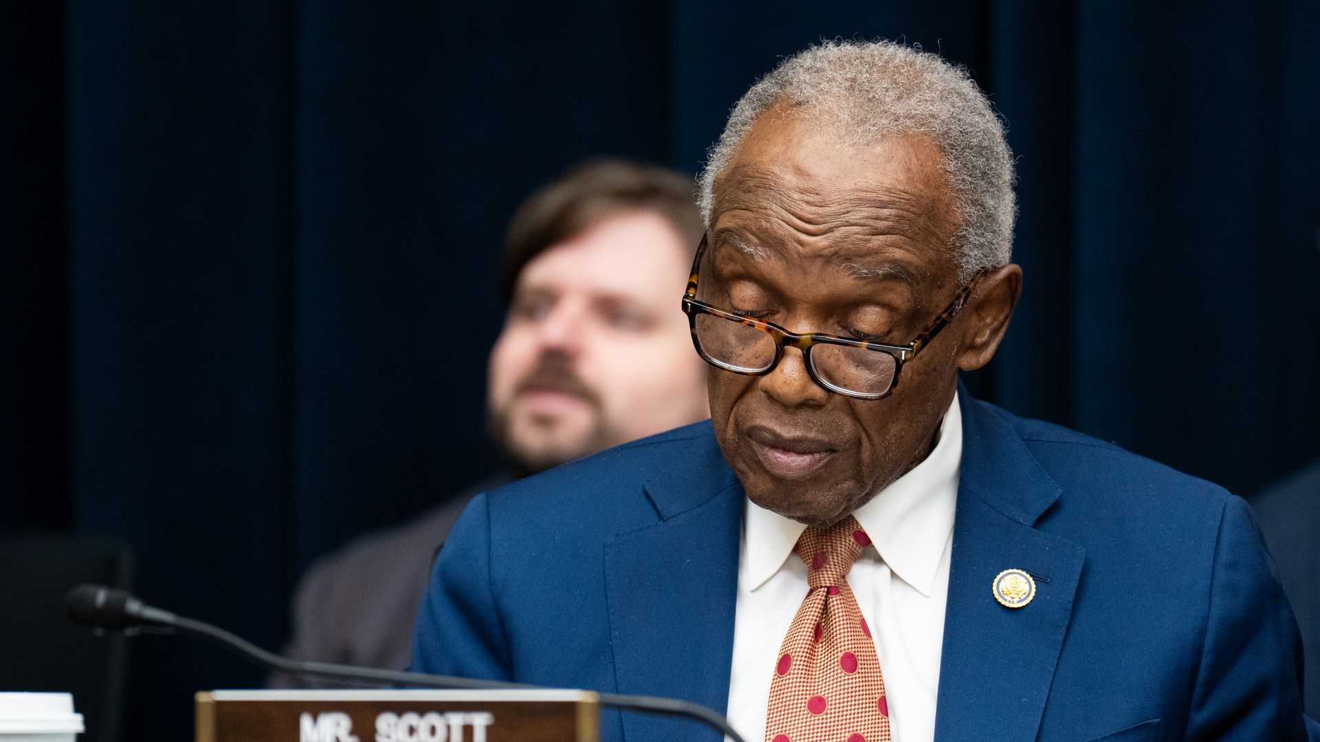 Close-up of an African American man with gray hair and glasses, in a blue suit and orange polka-dot tie, seated at a desk with a "MR. SCOTT" nameplate during a hearing.