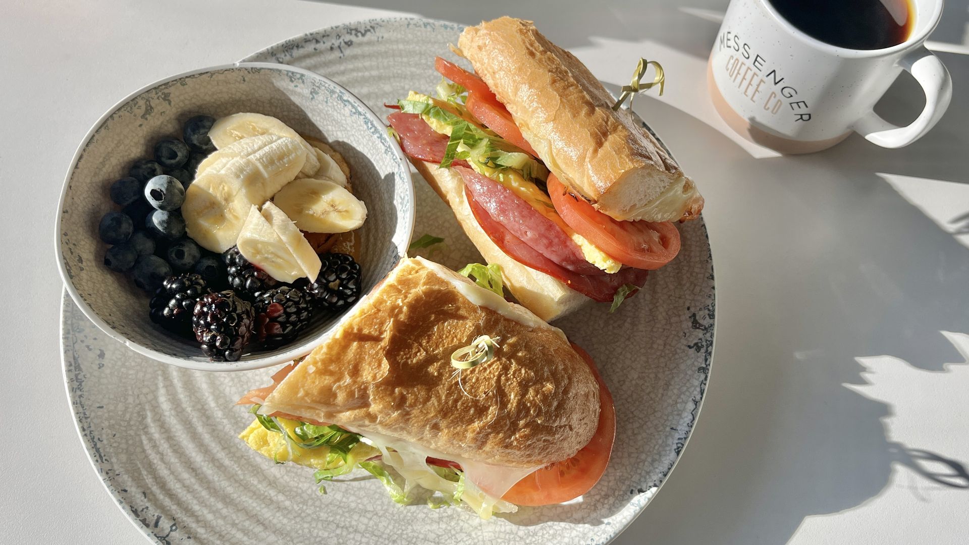 Plate with two sandwiches containing lettuce, tomato, egg, meat, and cheese, a bowl of banana slices, blueberries, and blackberries, and a white coffee cup with black coffee.