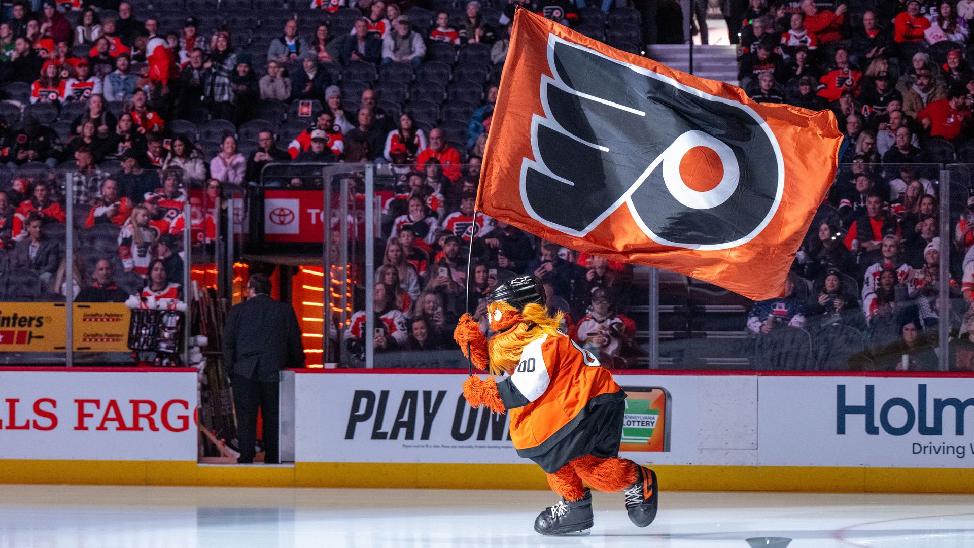 The Gritty orange mascot skating on an ice rink holding black and orange flag of the Flyers hockey team. 