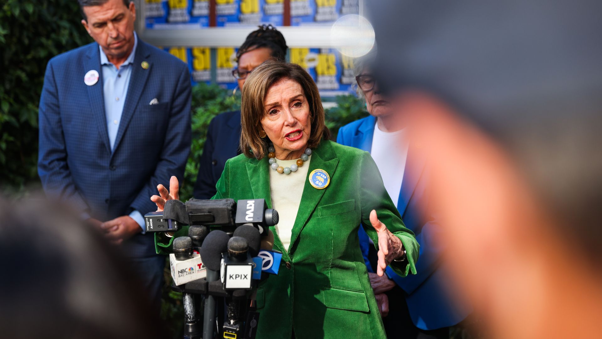 Nancy Pelosi, wearing a green suit jacket, speaks into a cluster of microphones while flanked by fellow House Democrats in front of a hedge and several blue posters.