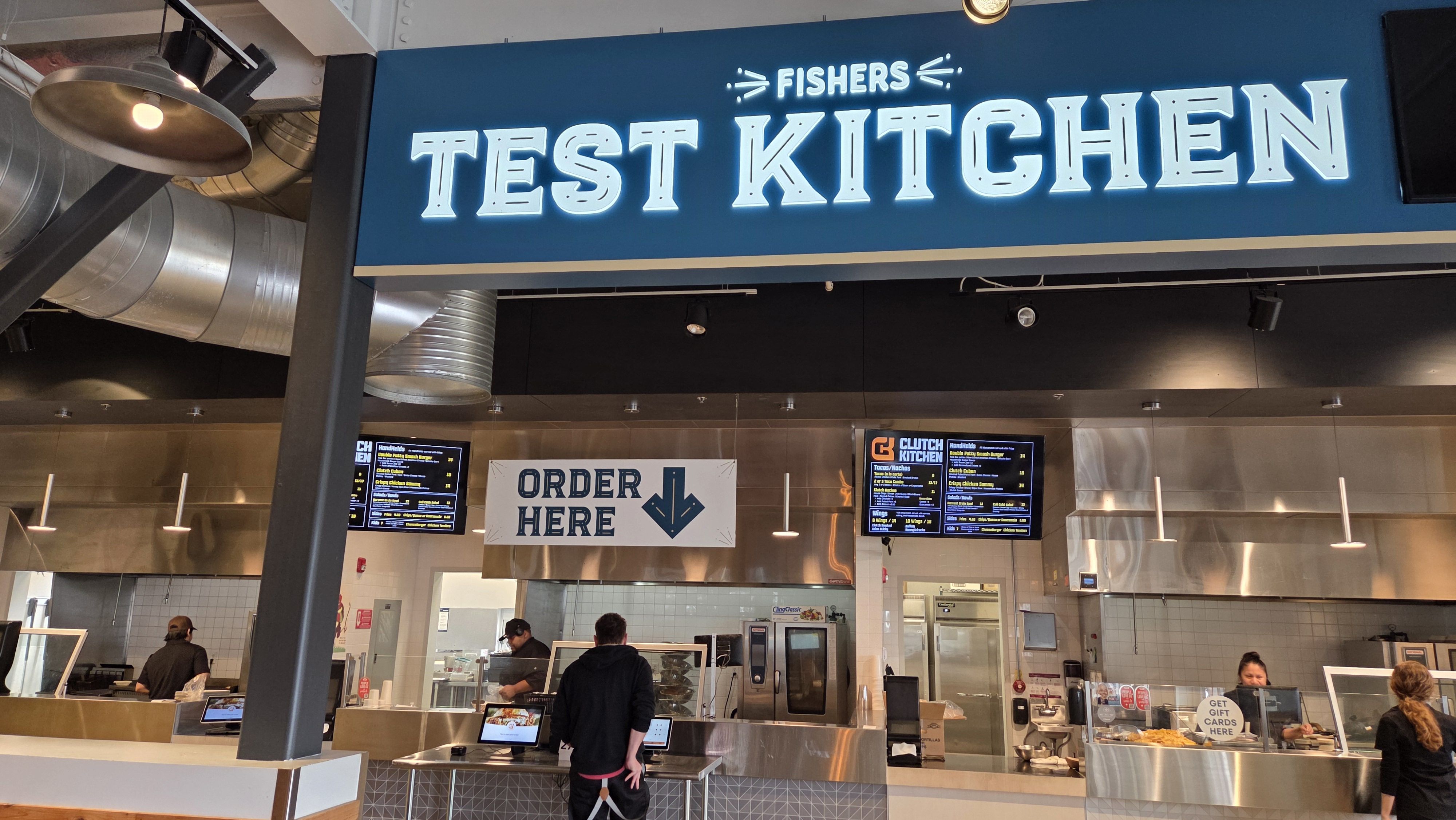 Interior of Fishers Test Kitchen with a large blue sign, a white 'Order Here' sign, digital menu boards, staff preparing food behind counters, and visible kitchen equipment.
