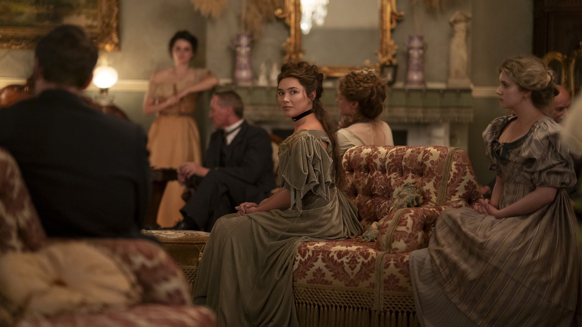 A young woman played by Florence Pugh, sits in a green-gray gown on a patterned sofa in the center of a parlor scene.