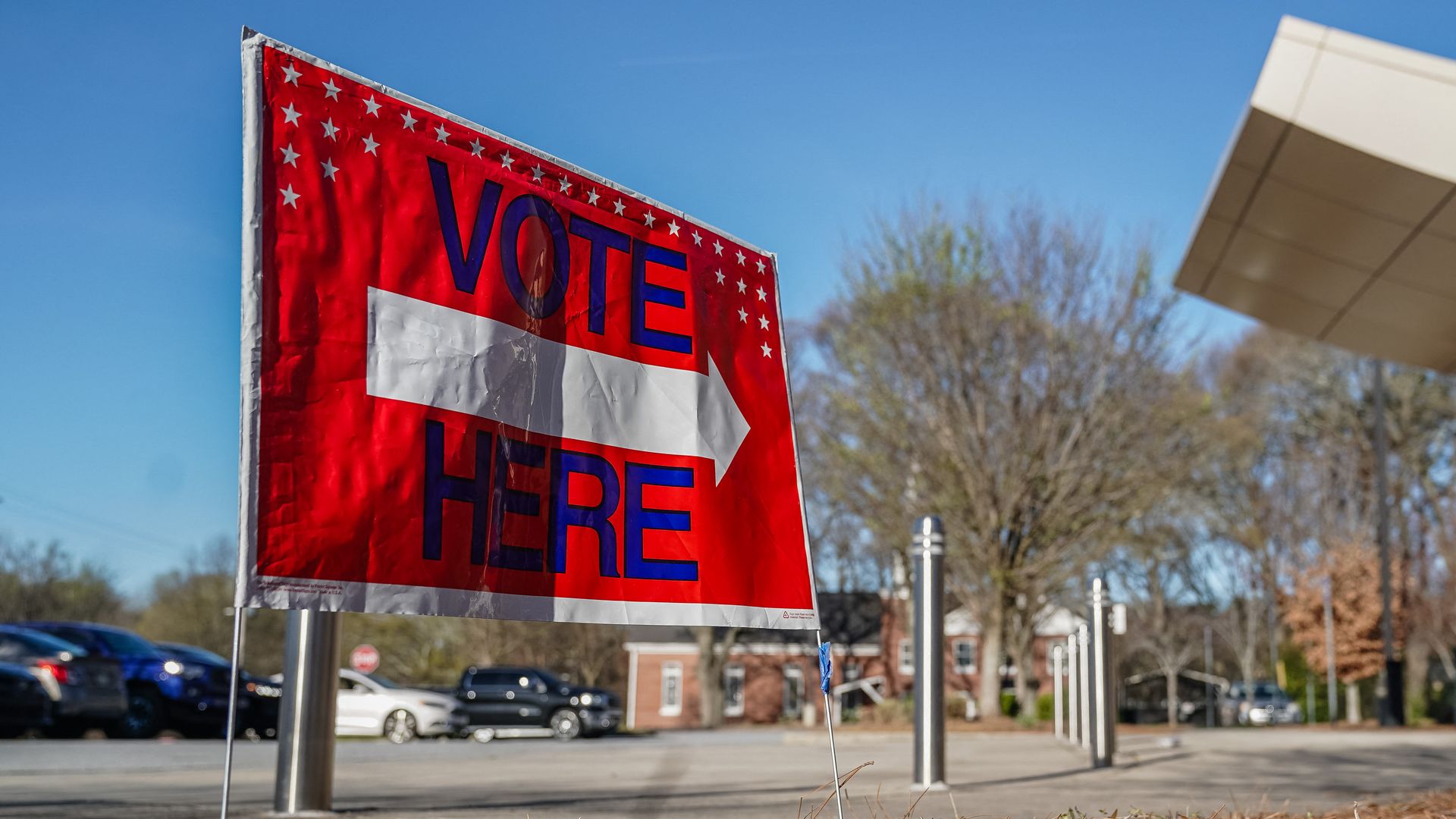 A red sign with a white arrow pointing to the right and "Vote Here" written in blue, with small white stars on upper half of the sign.