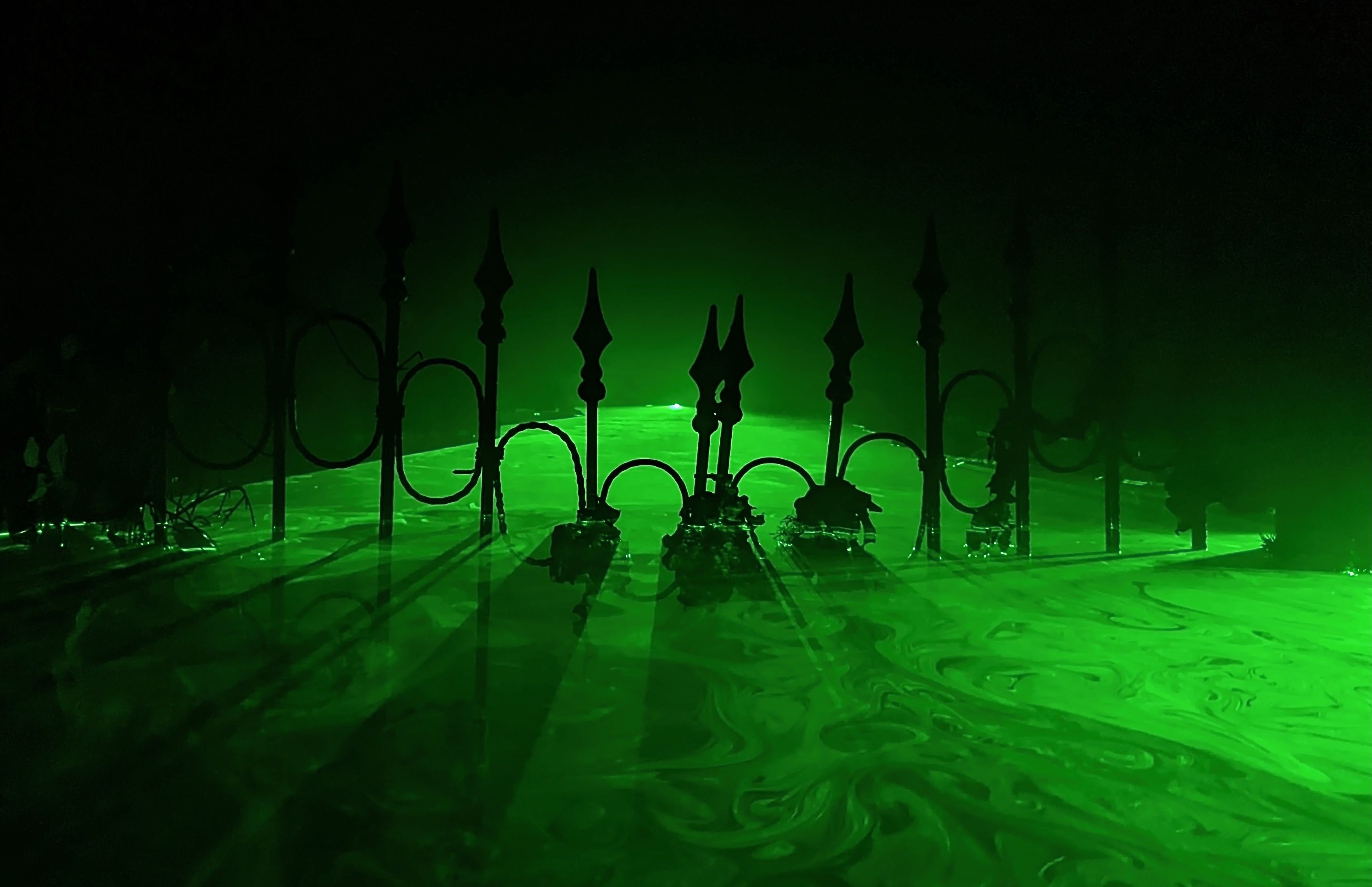 A green laser shines over fog to make a layer of light that resembles a swamp around a wrought iron fence. 