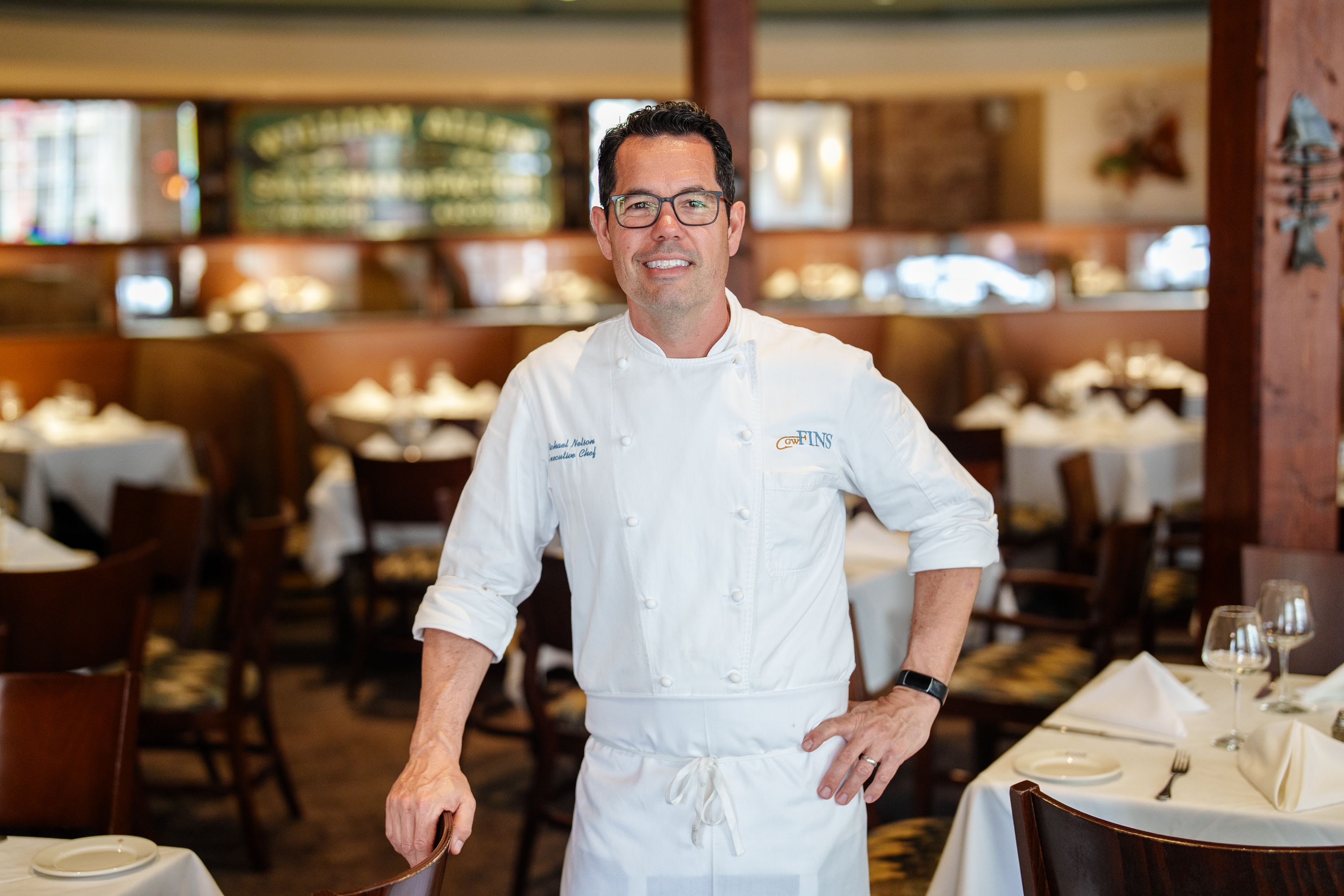 Chef Mike Nelson, wearing his chef's coat, smiles in a posed photo inside an empty restaurant.
