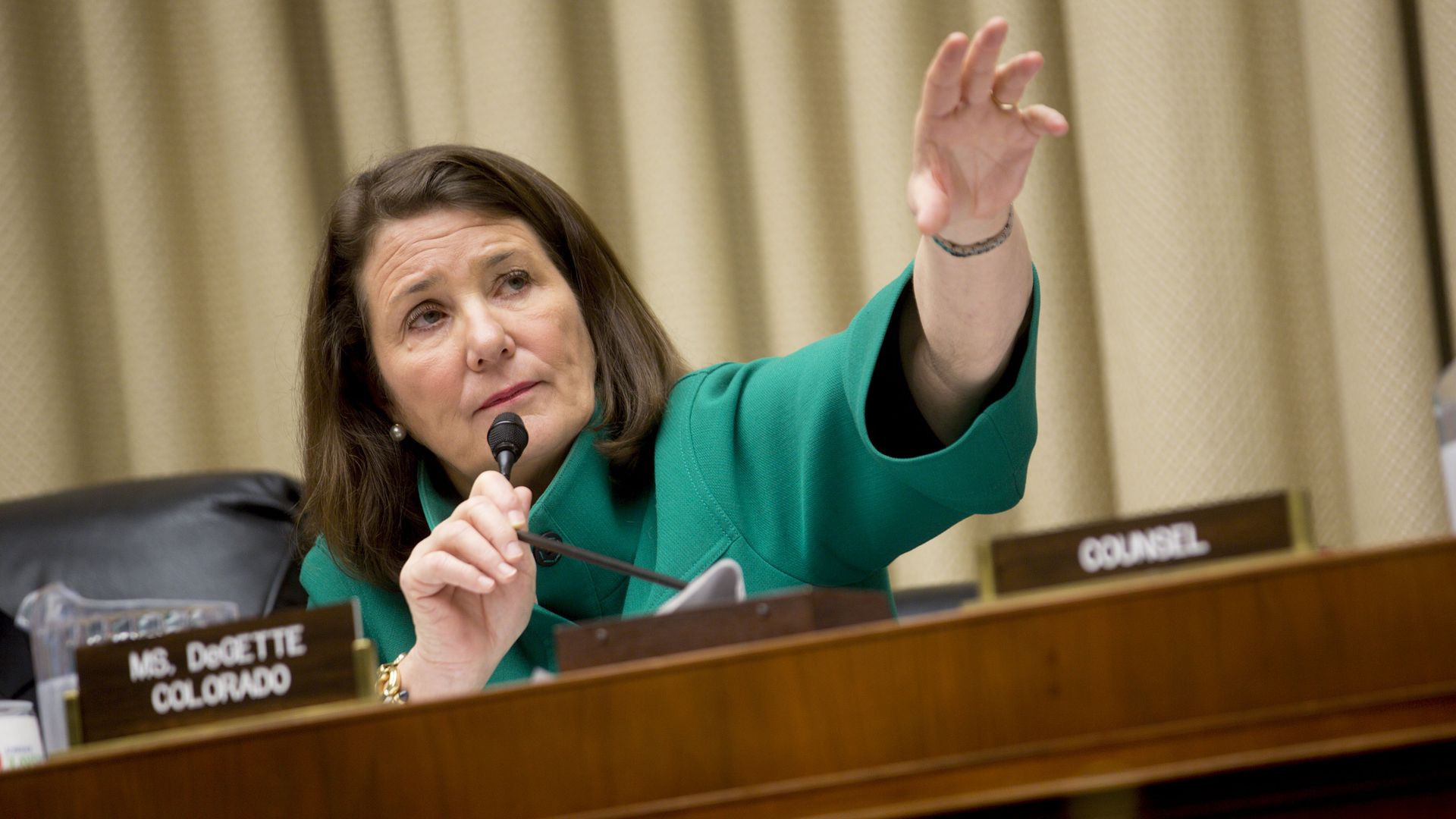 A woman in a teal blazer sits at a council desk, speaks into a microphone, and raises her right hand. Beige curtains and nameplates are visible behind her.