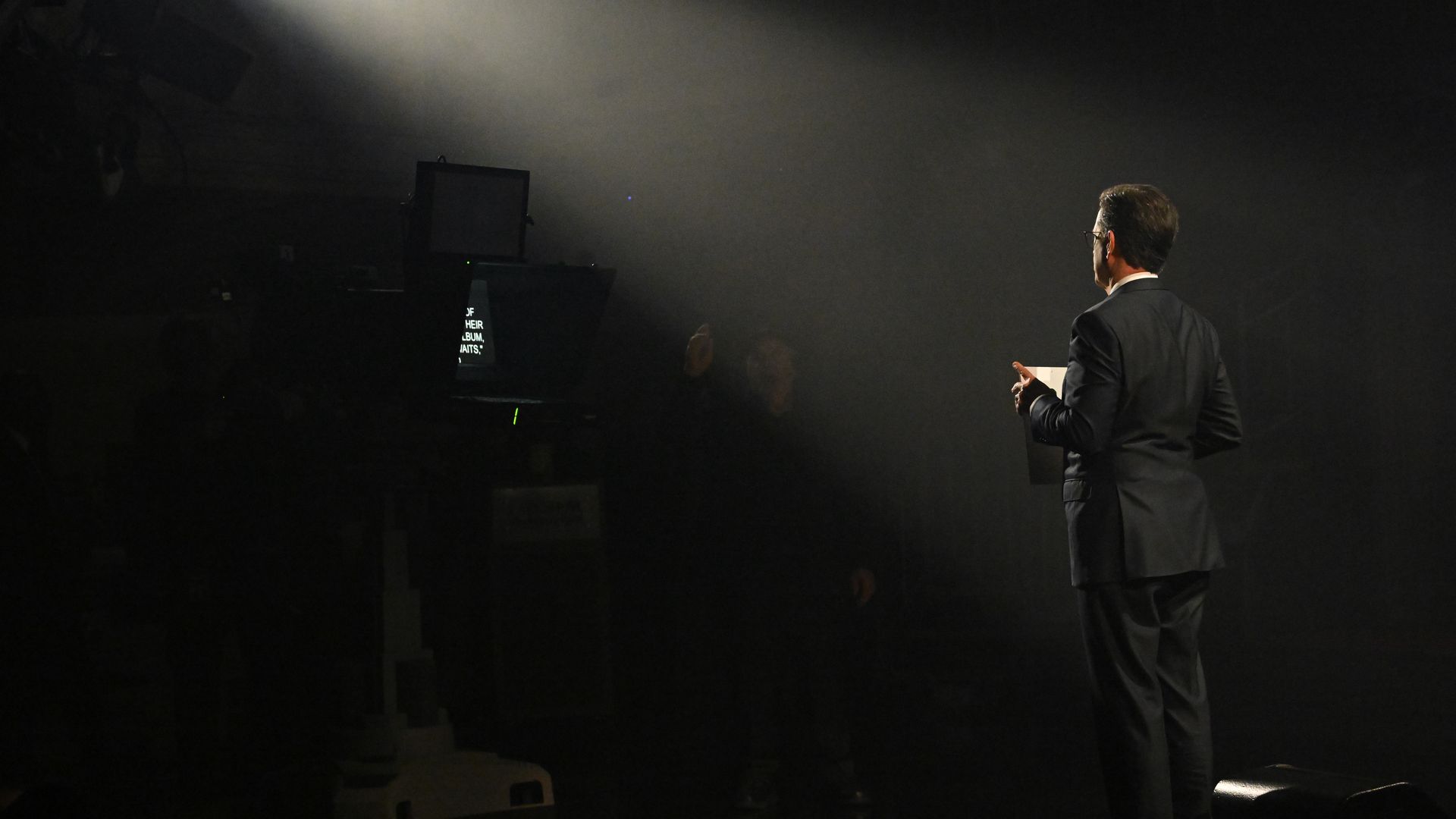 Man in a dark suit stands on a dim stage, seen from the back, as a strong spotlight from the left pierces the fog, lighting his silhouette on the right.