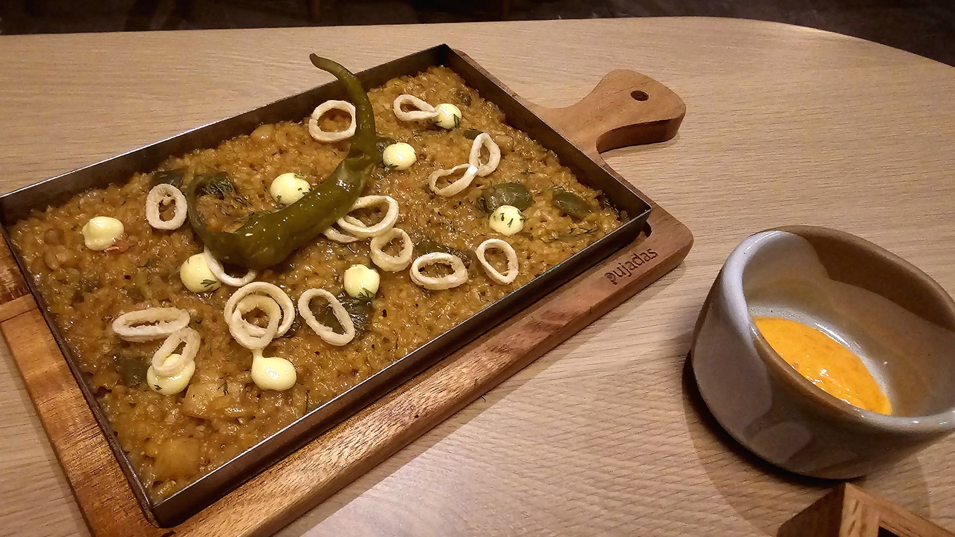 Rectangular metal pan of orange-spiced rice topped with a roasted green chili, cuttlefish, and small white dollops; placed on a wooden board beside a ceramic sauce boat with orange sauce.