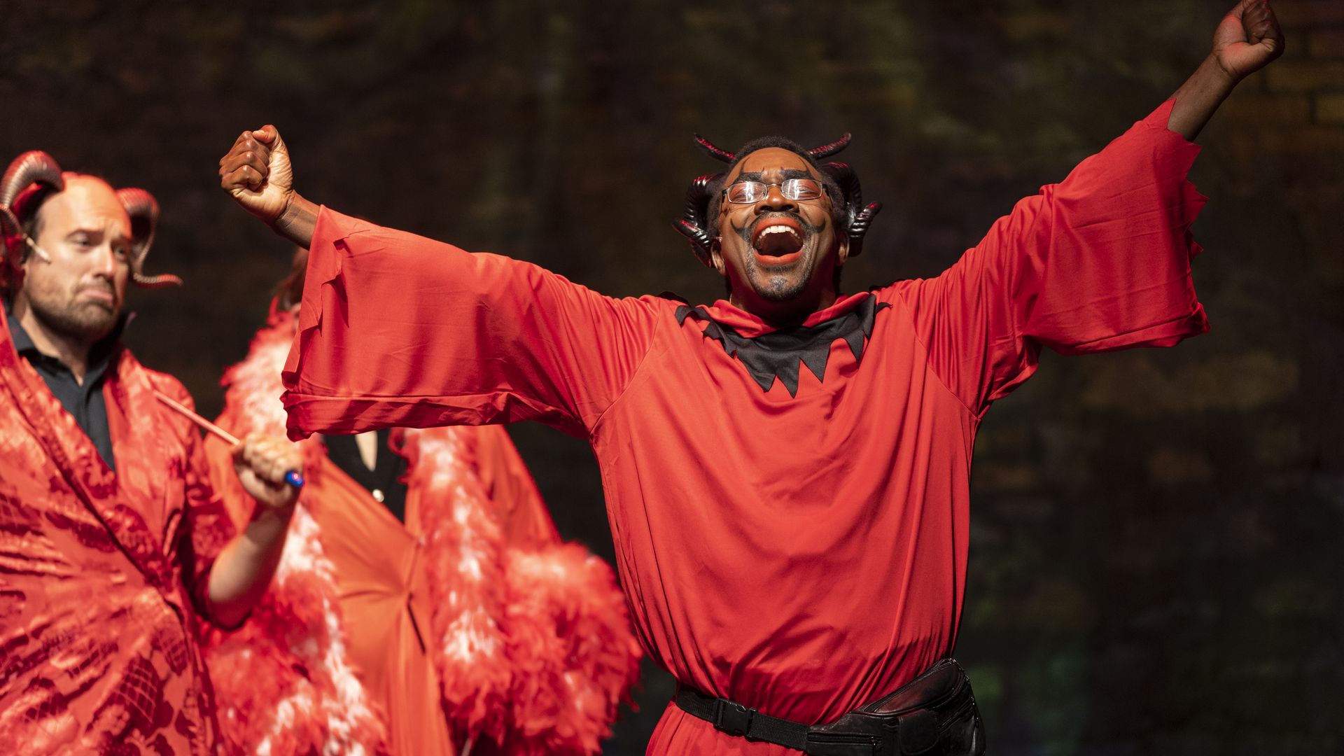 A man dressed as the devil stretches out his arms while singing.