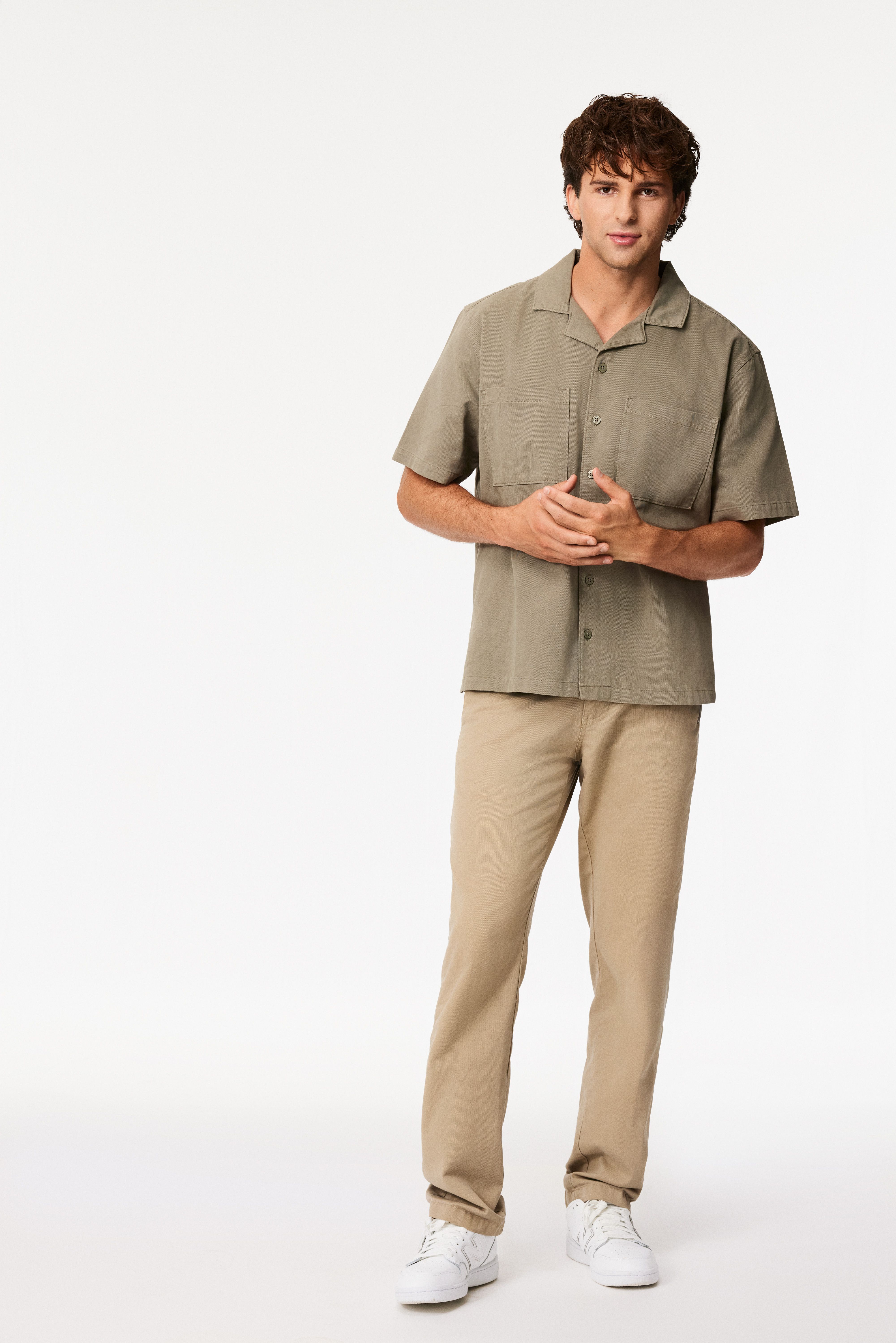 Young man with curly brown hair wearing a khaki short-sleeve button-up shirt, beige pants, and white sneakers standing against a white background.