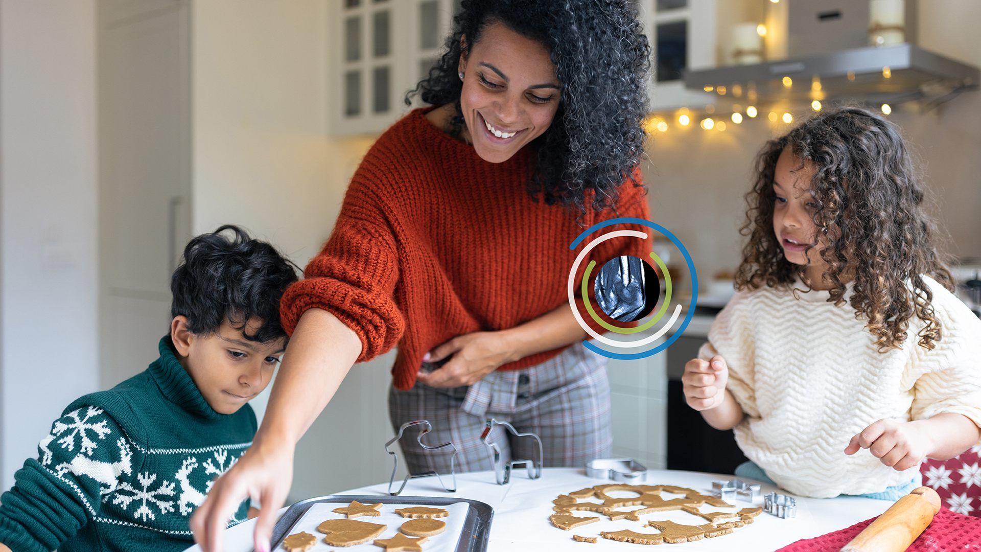 Smiling woman in a red knit sweater guides two children as they bake cookies in a bright kitchen, using cutters on a tray with warm fairy lights in the background.