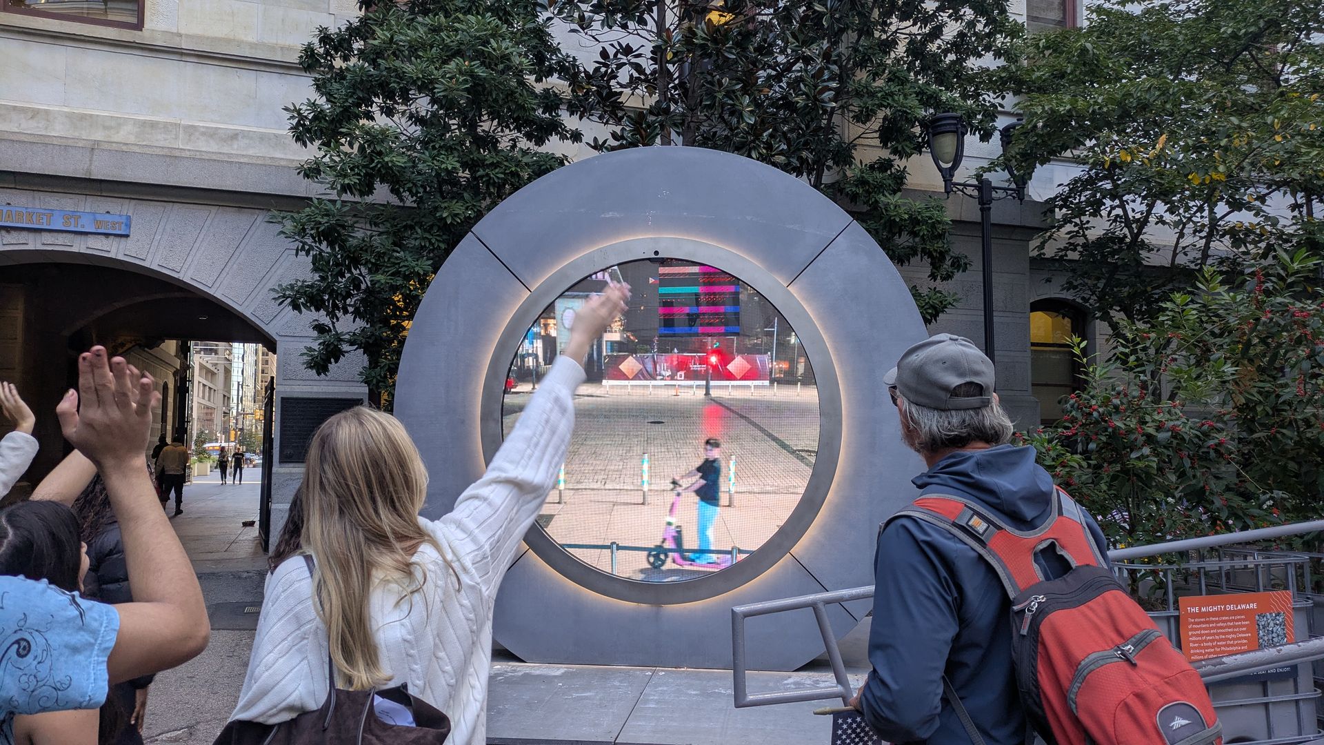 People stand around a large circular metallic sculpture with a glowing ring and inner screen; a hand waves through the opening as a city street and a Market St. West sign appear behind.