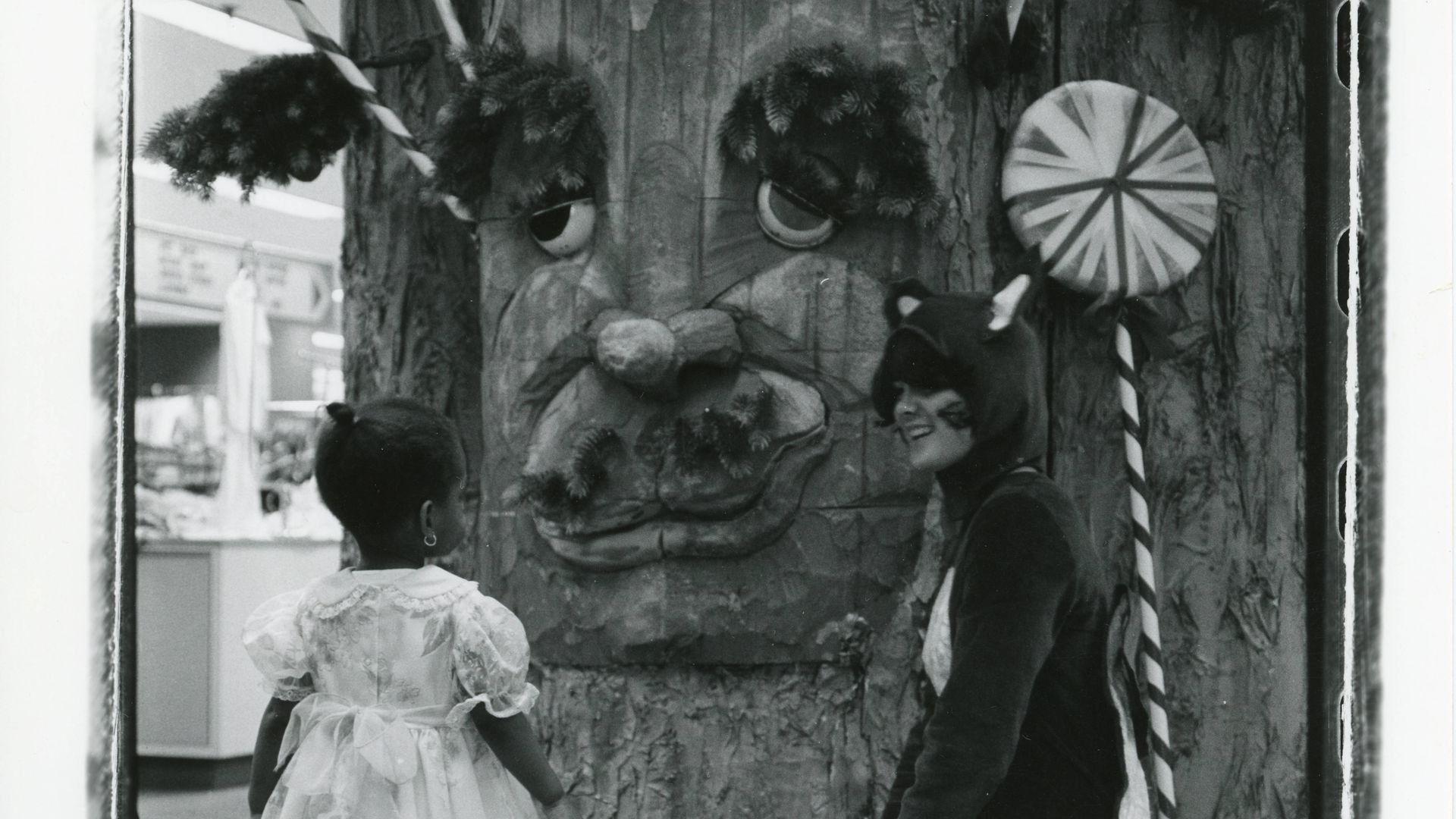 A historic black-and-white photo of a child speaking with "Mr. Tree," a massive talking tree puppet, alongside a woman in a fox onesie