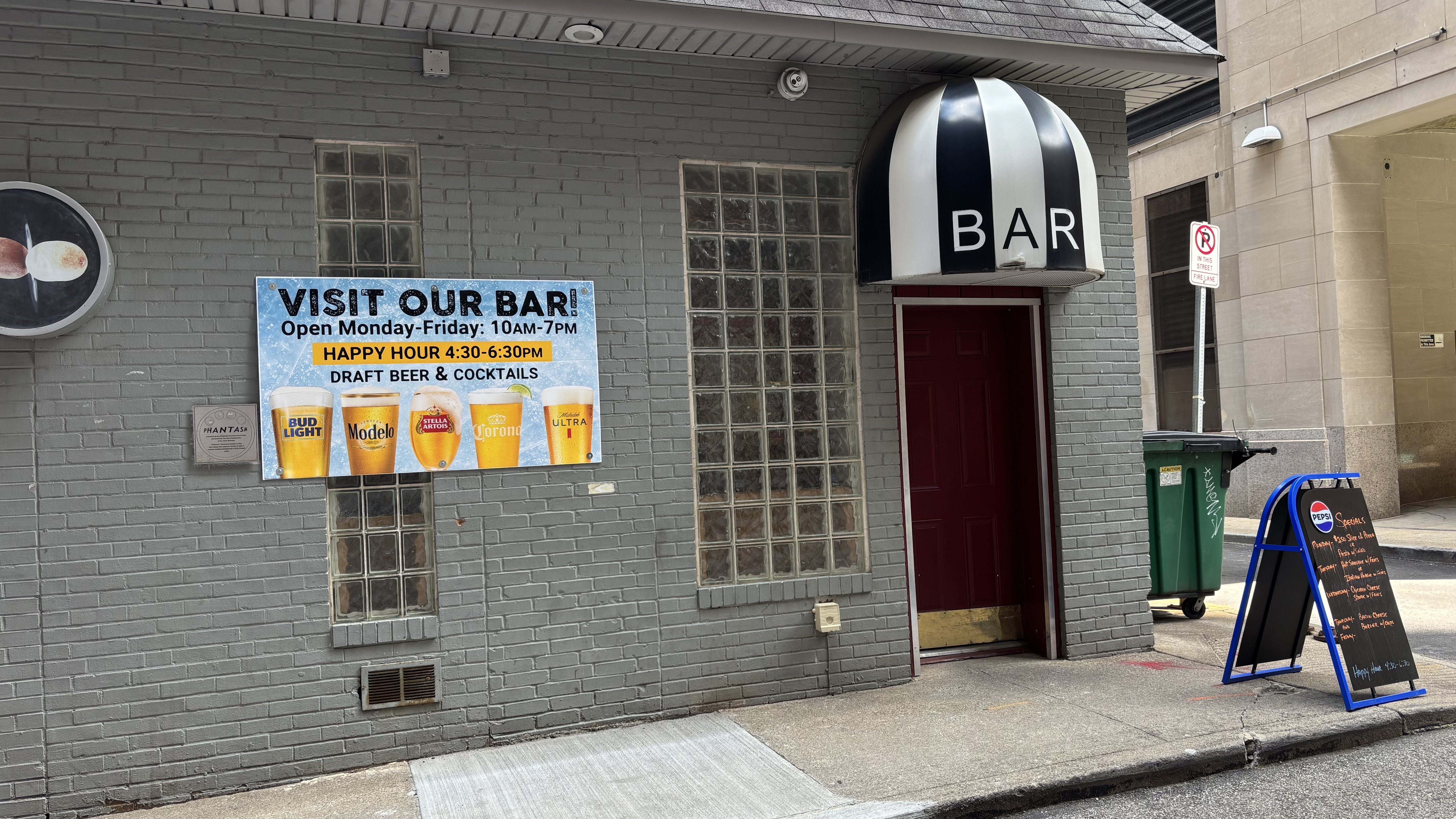Gray brick building with a striped black-and-white dome sign reading BAR over a red door. A blue "VISIT OUR BAR" sign advertises beer and happy hour; glass block windows flank the entrance.