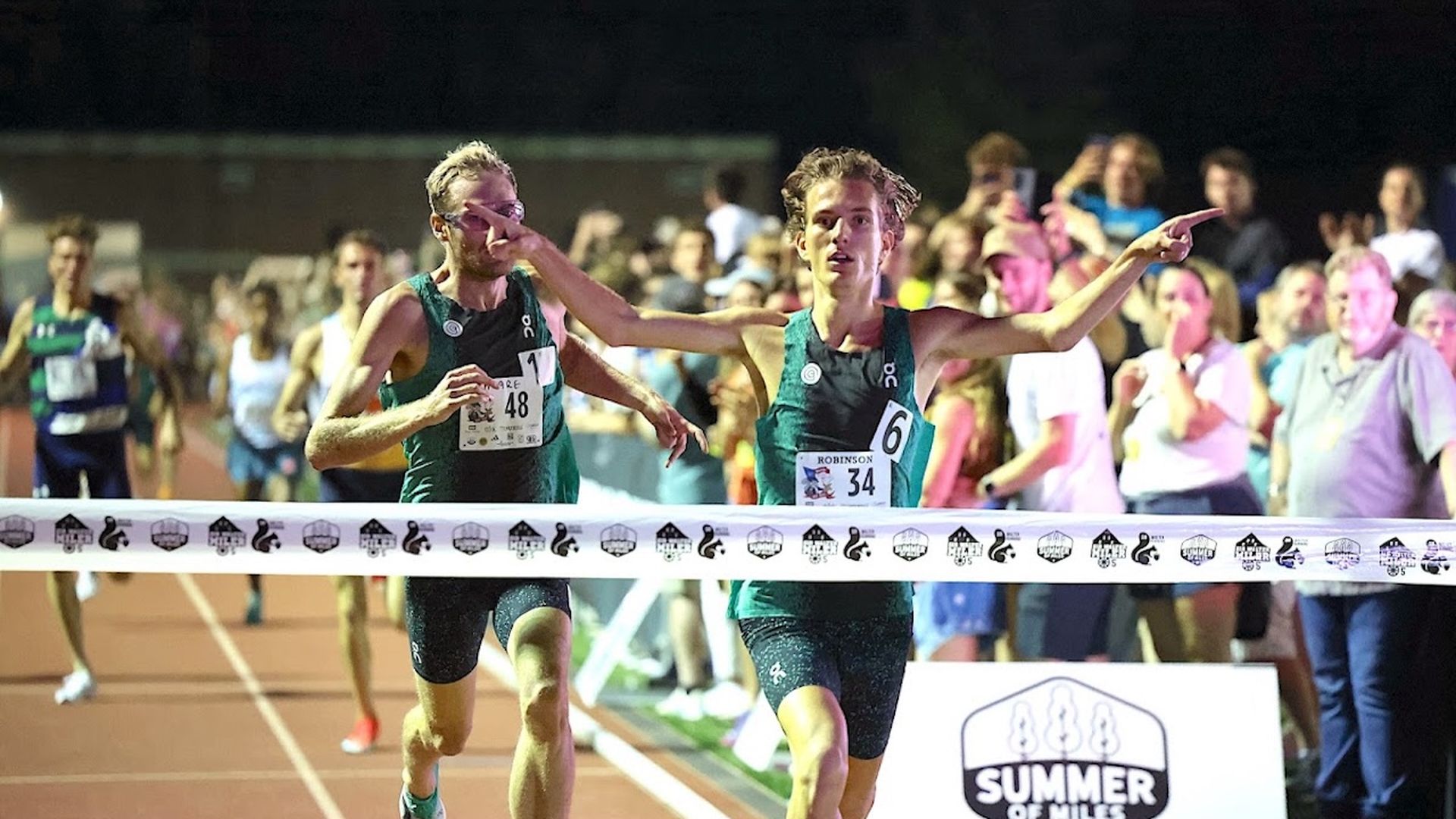 Two male runners in green uniforms cross the track finish line, one pointing ahead while the other shields his face; a cheering crowd and the "Summer of Miles" banner in the background.
