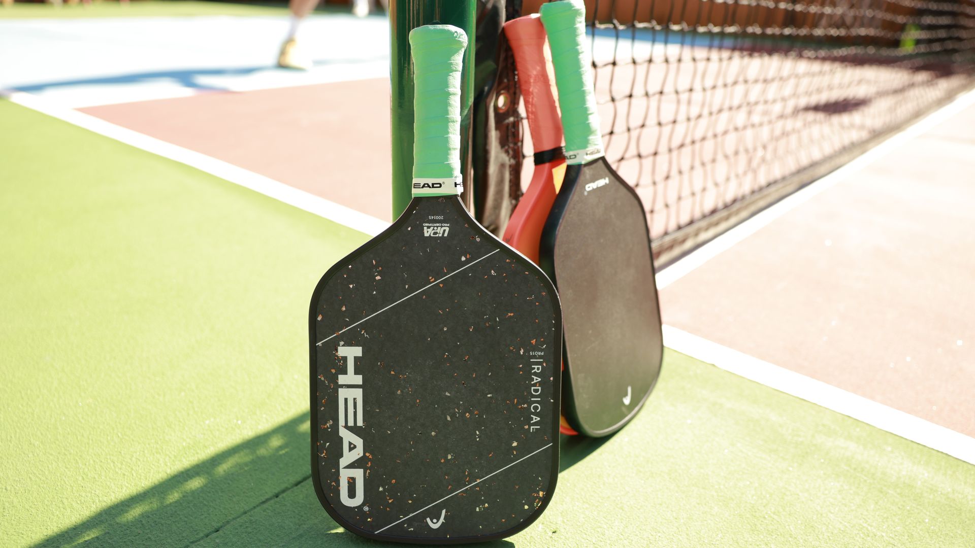 Several pickleball paddles with green grips leaning against a net post on a bright outdoor court. Green synthetic surface with white boundary lines, a mesh net to the right, and a blurred player in the background.