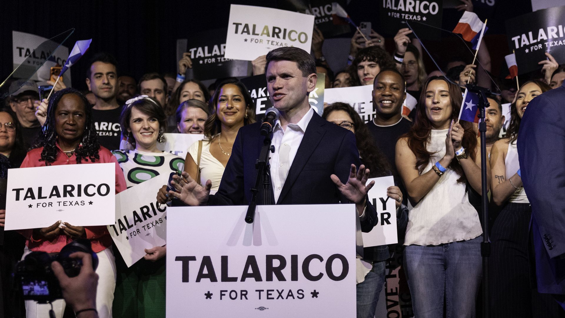 A man in a dark suit speaks at a podium reading "TALARICO FOR TEXAS" as a cheering crowd behind him holds signs reading "TALARICO FOR TEXAS" and waves small Texas flags.